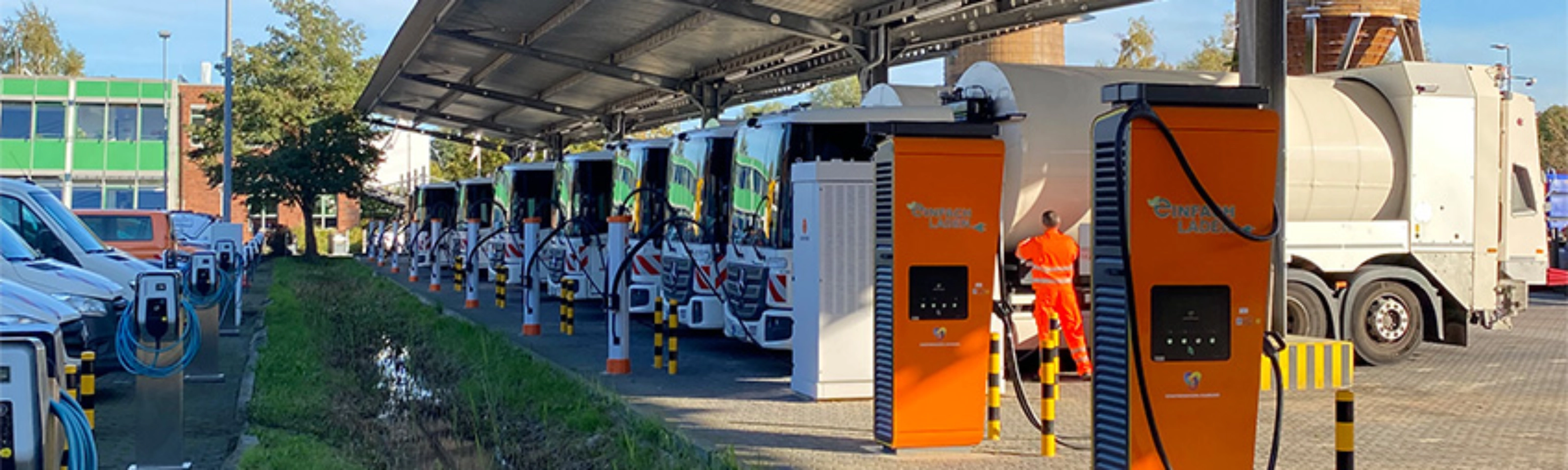 Orange charging stations beneath a solar canopy at a parking area with parked trucks.