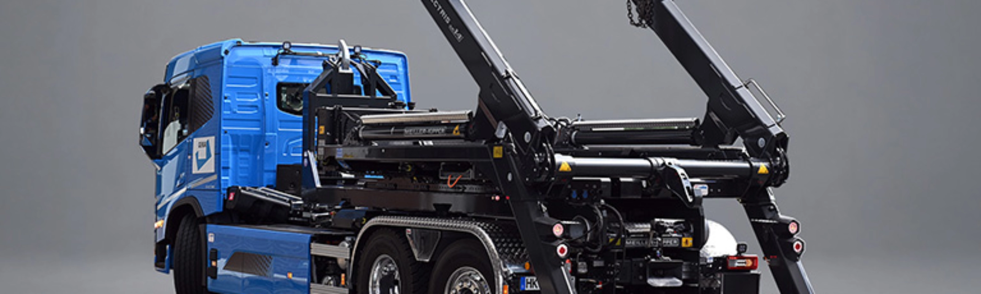 Blauer Lkw mit schwarzem Meiller-Hakenliftsystem und angehobenen Hubarmen in schräger Rückansicht.
