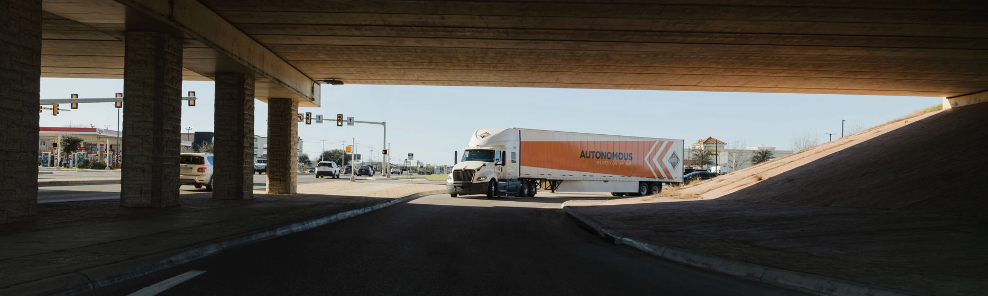 A self-driving truck with an "Autonomous" trailer drives through an intersection under a bridge.