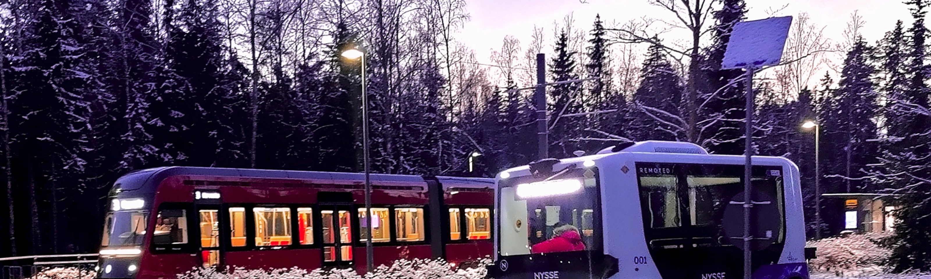 Autonomous shuttle bus driving beside a tram on a snowy street.