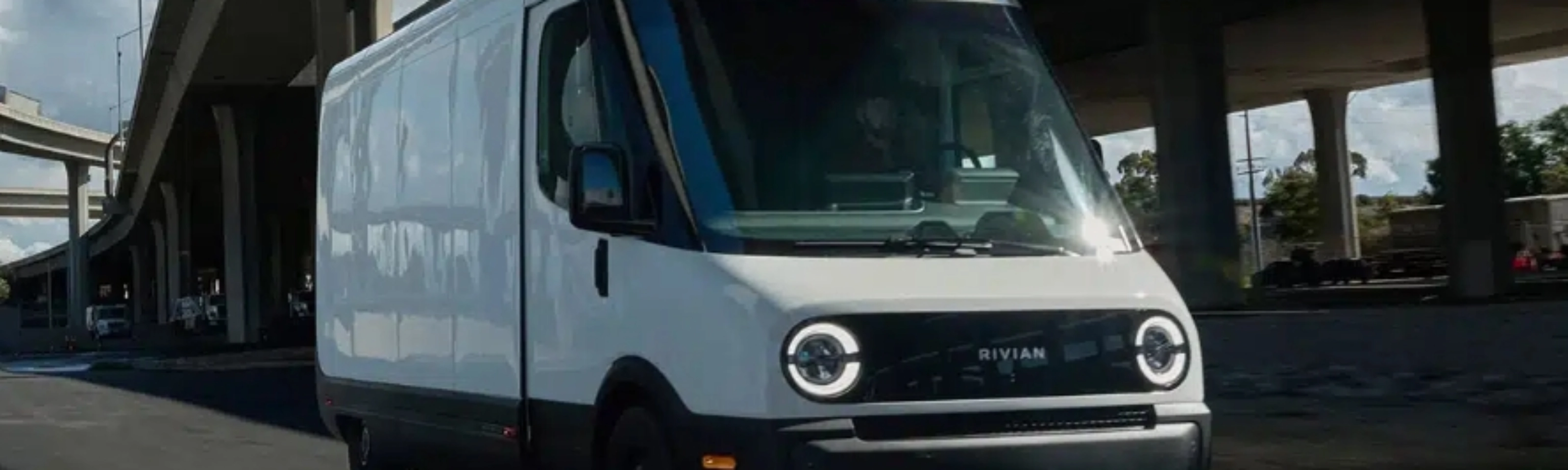 White Rivian electric delivery van driving on a city street under a highway bridge.