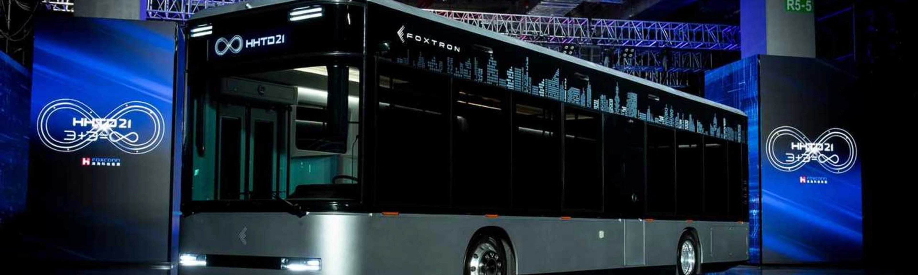 A modern electric city bus is showcased indoors with the logo "Foxtron" and digital skyline.