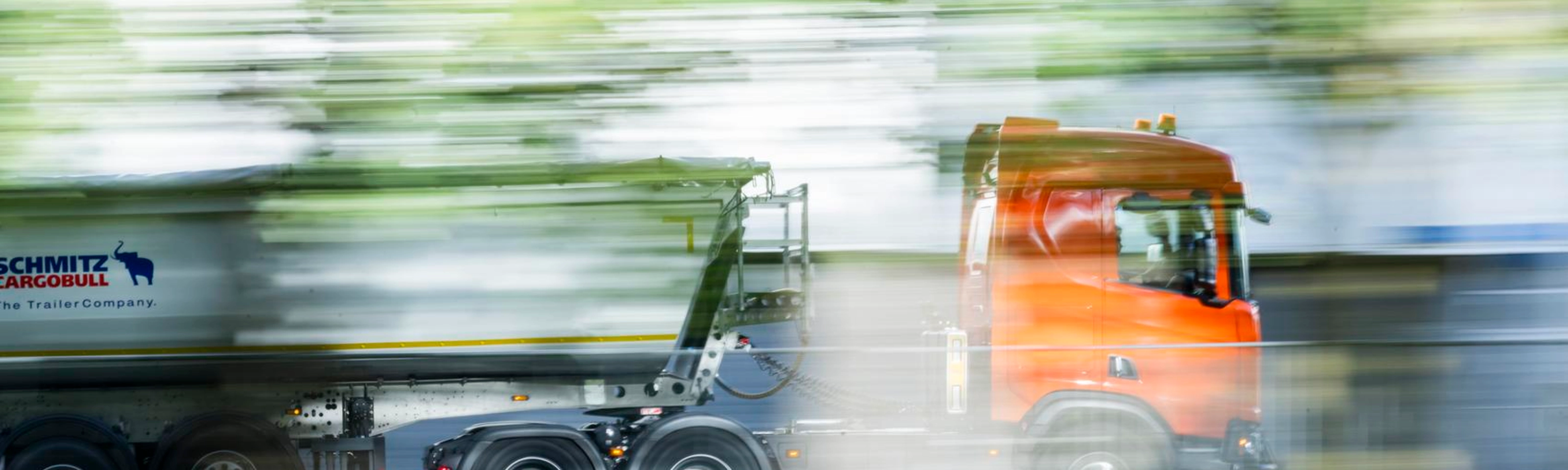 A truck with a trailer drives along a road surrounded by greenery.