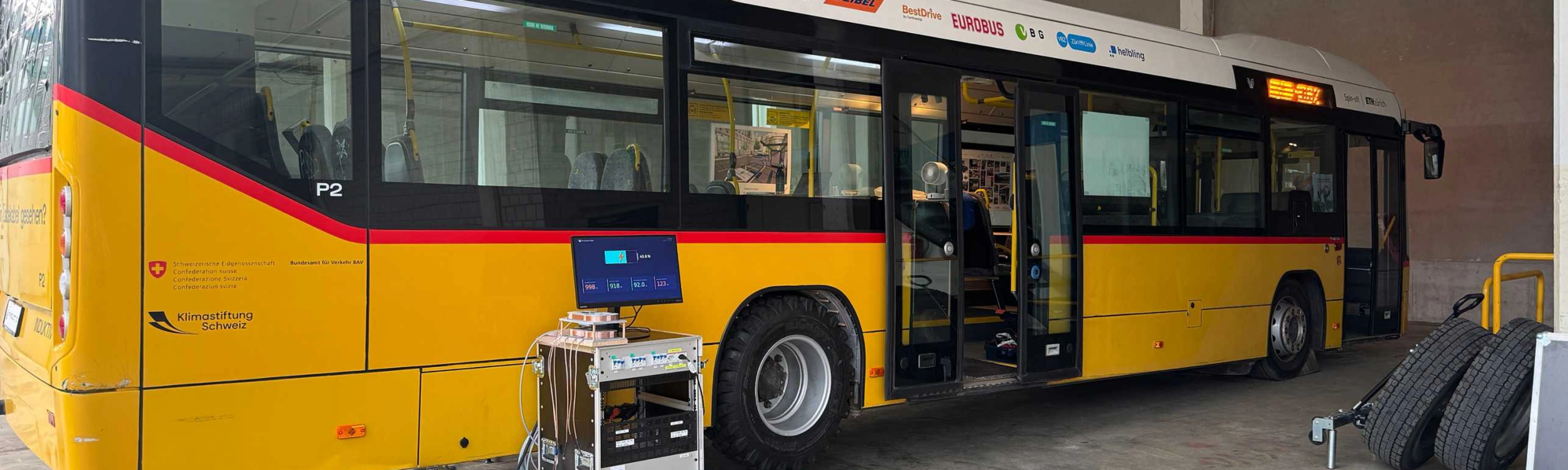 Yellow electric bus in a workshop with open doors and technical equipment in front of it.