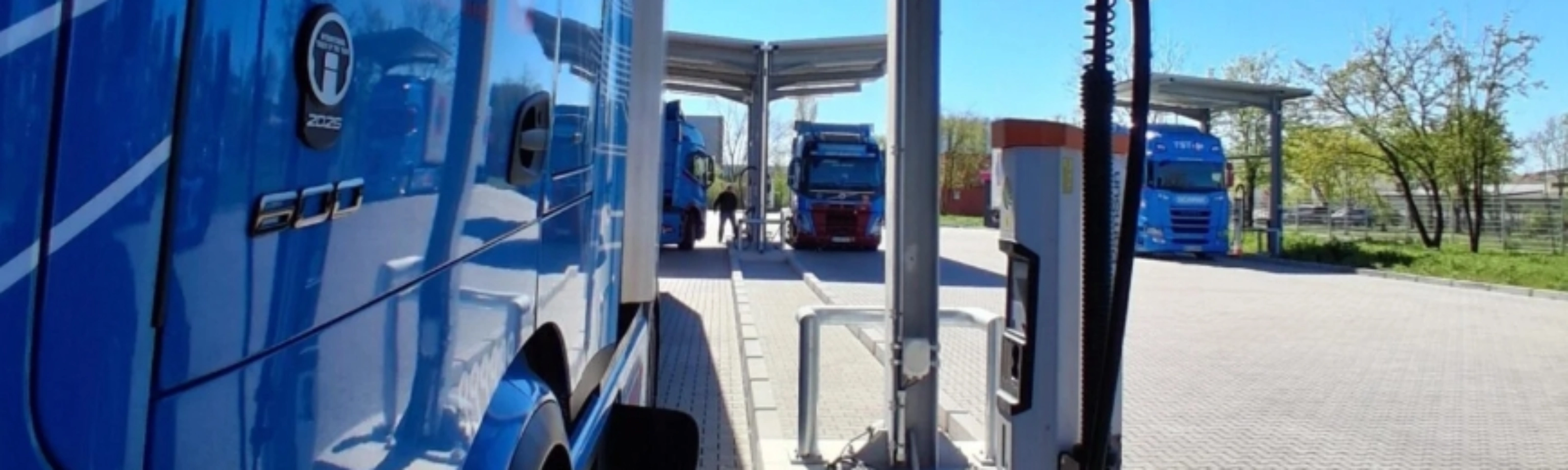 Blue electric truck connected to a charging station at a parking area.