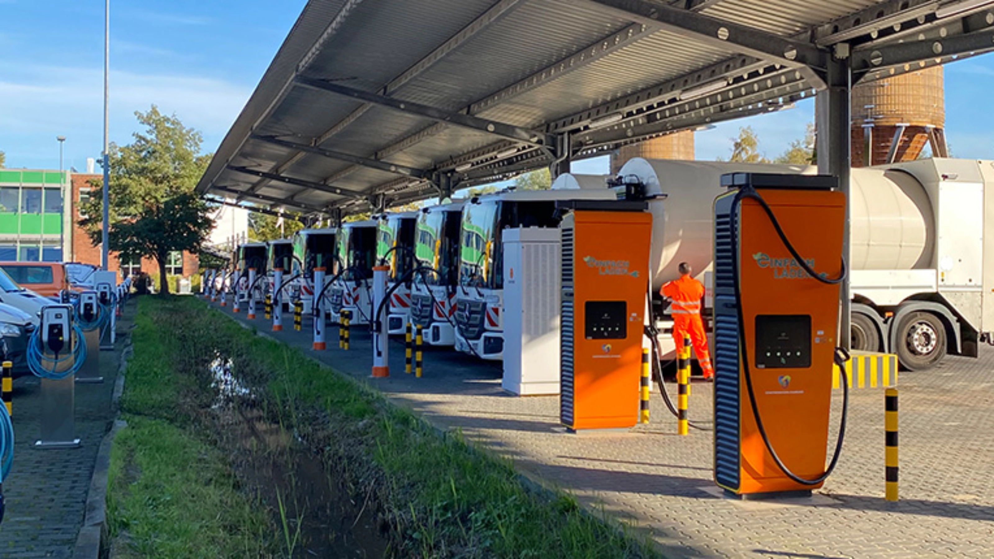Orange charging stations beneath a solar canopy at a parking area with parked trucks.