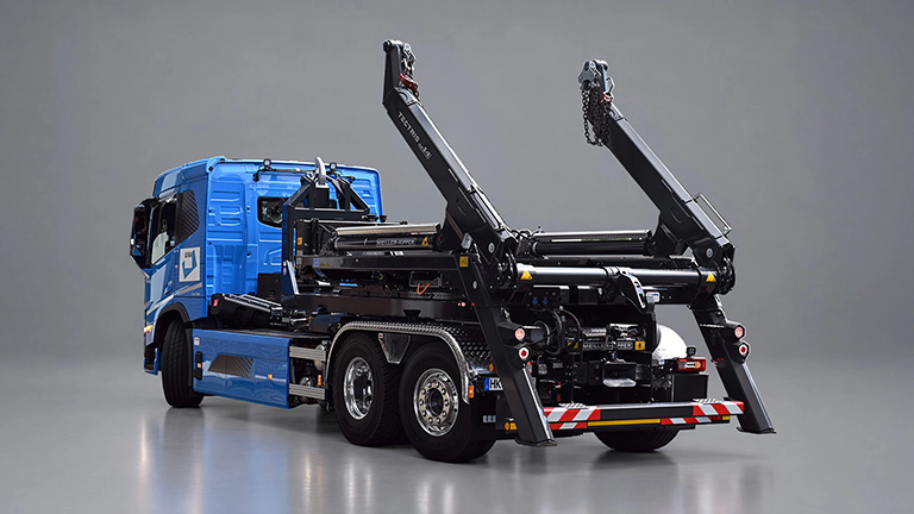Blauer Lkw mit schwarzem Meiller-Hakenliftsystem und angehobenen Hubarmen in schräger Rückansicht.