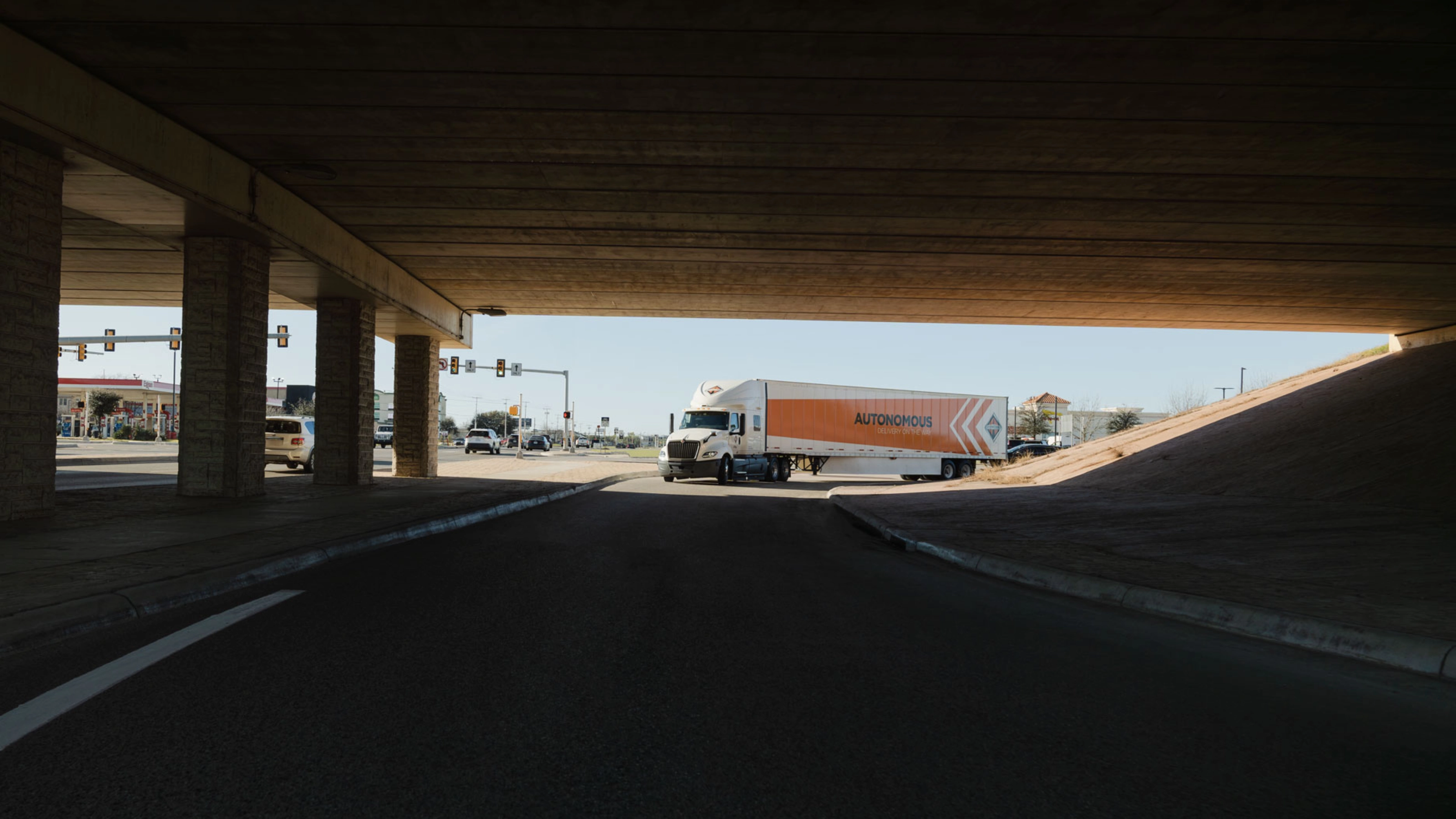 A self-driving truck with an "Autonomous" trailer drives through an intersection under a bridge.