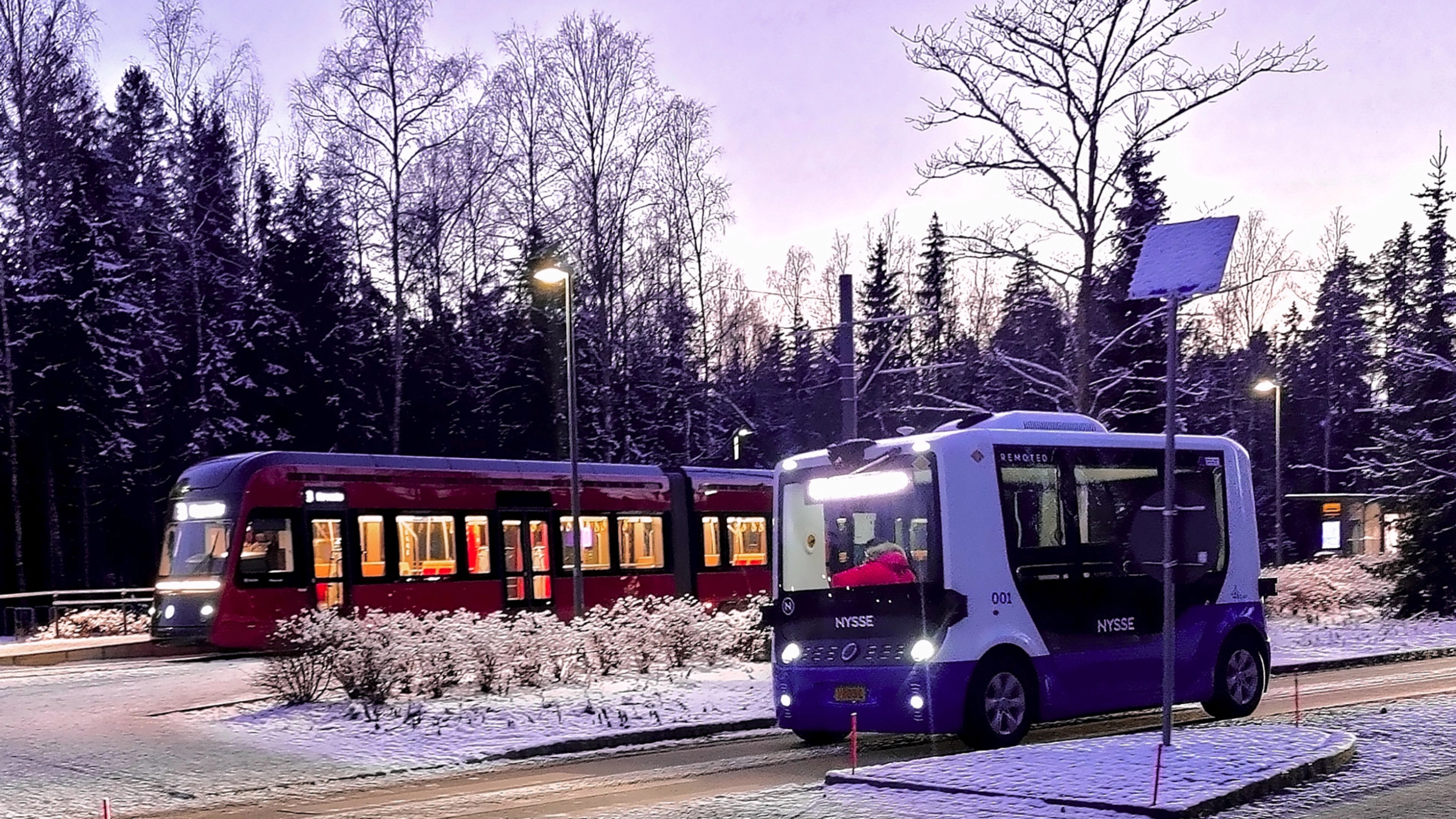 Autonomous shuttle bus driving beside a tram on a snowy street.