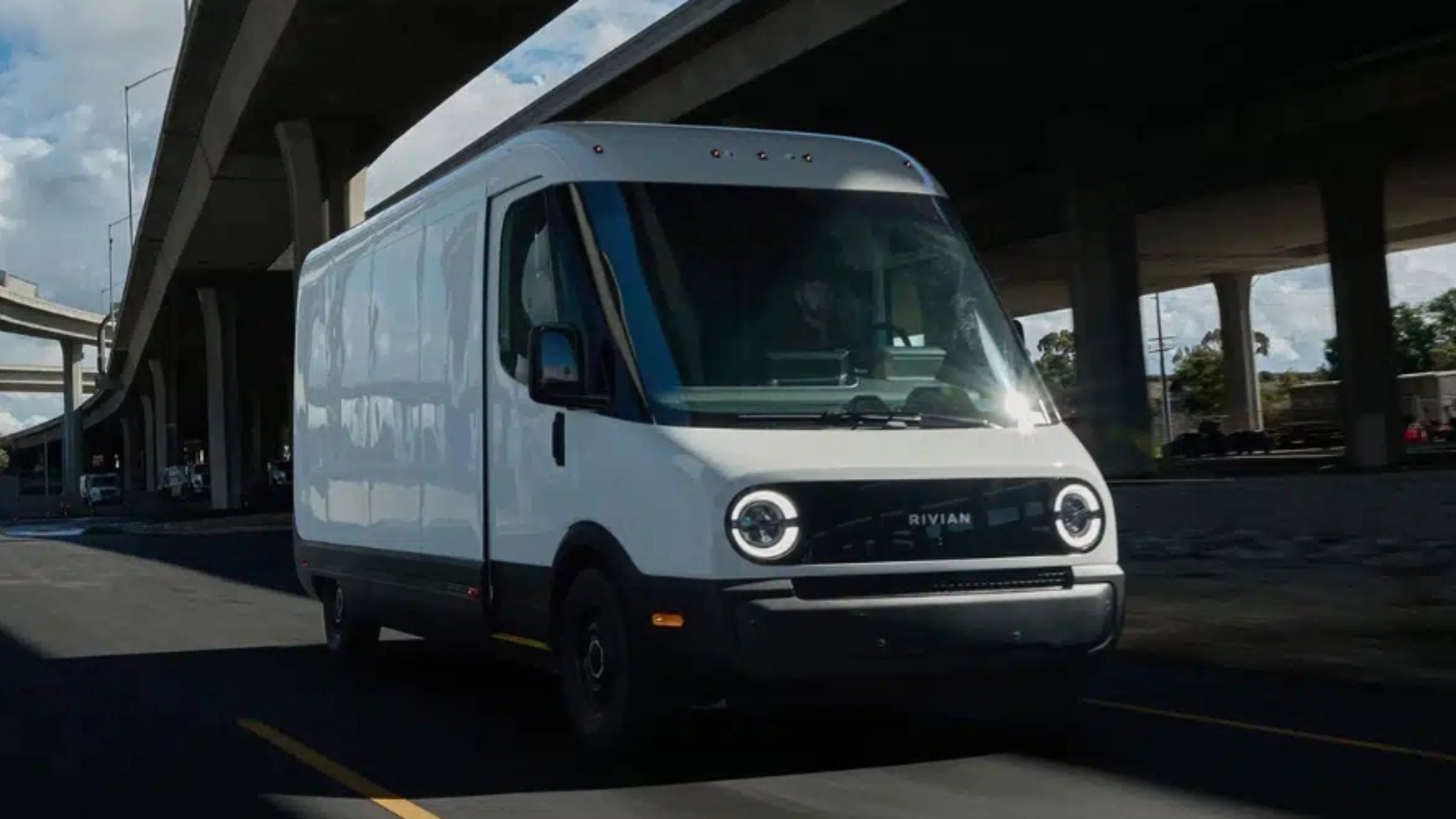 White Rivian electric delivery van driving on a city street under a highway bridge.