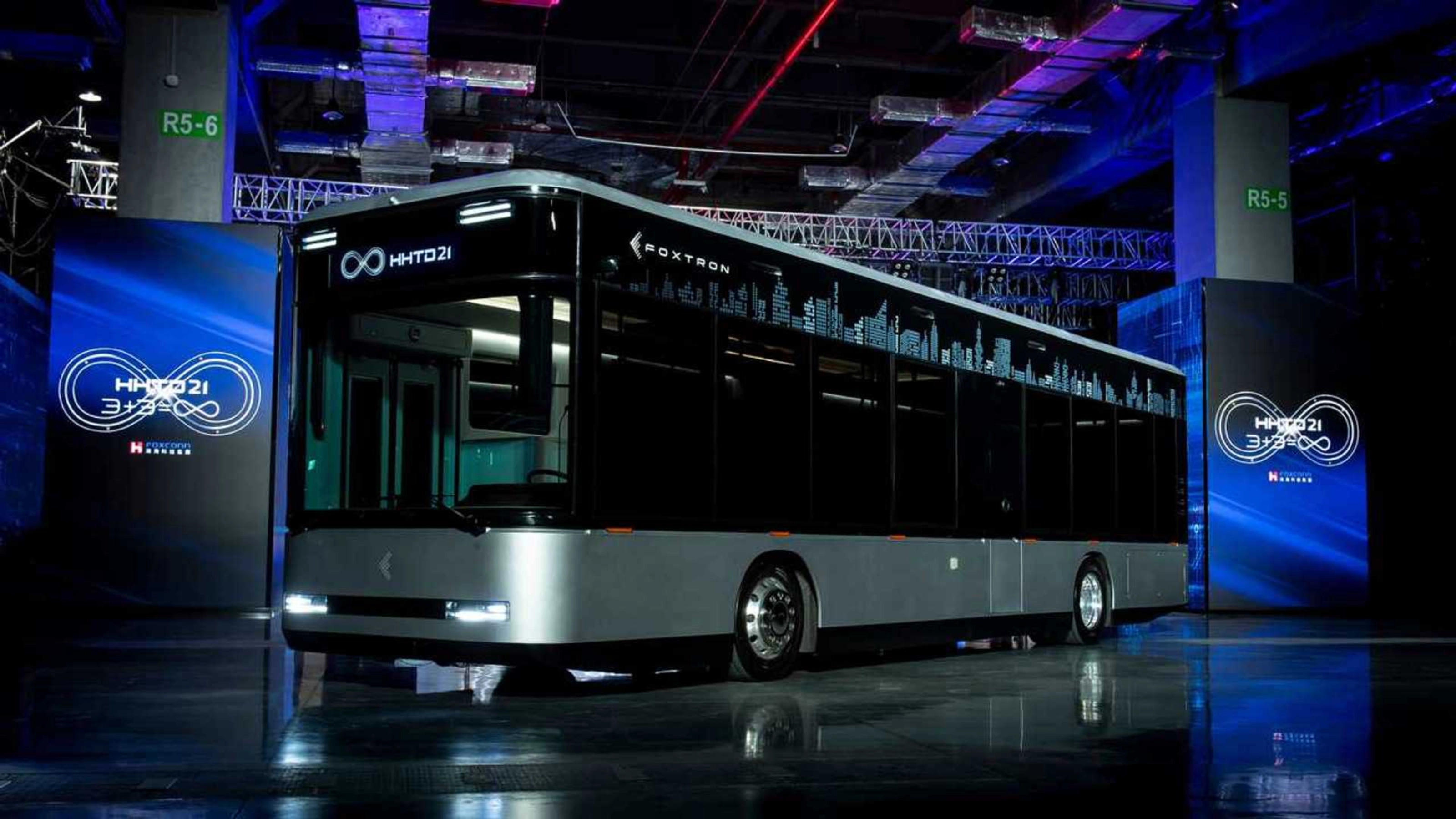A modern electric city bus is showcased indoors with the logo "Foxtron" and digital skyline.