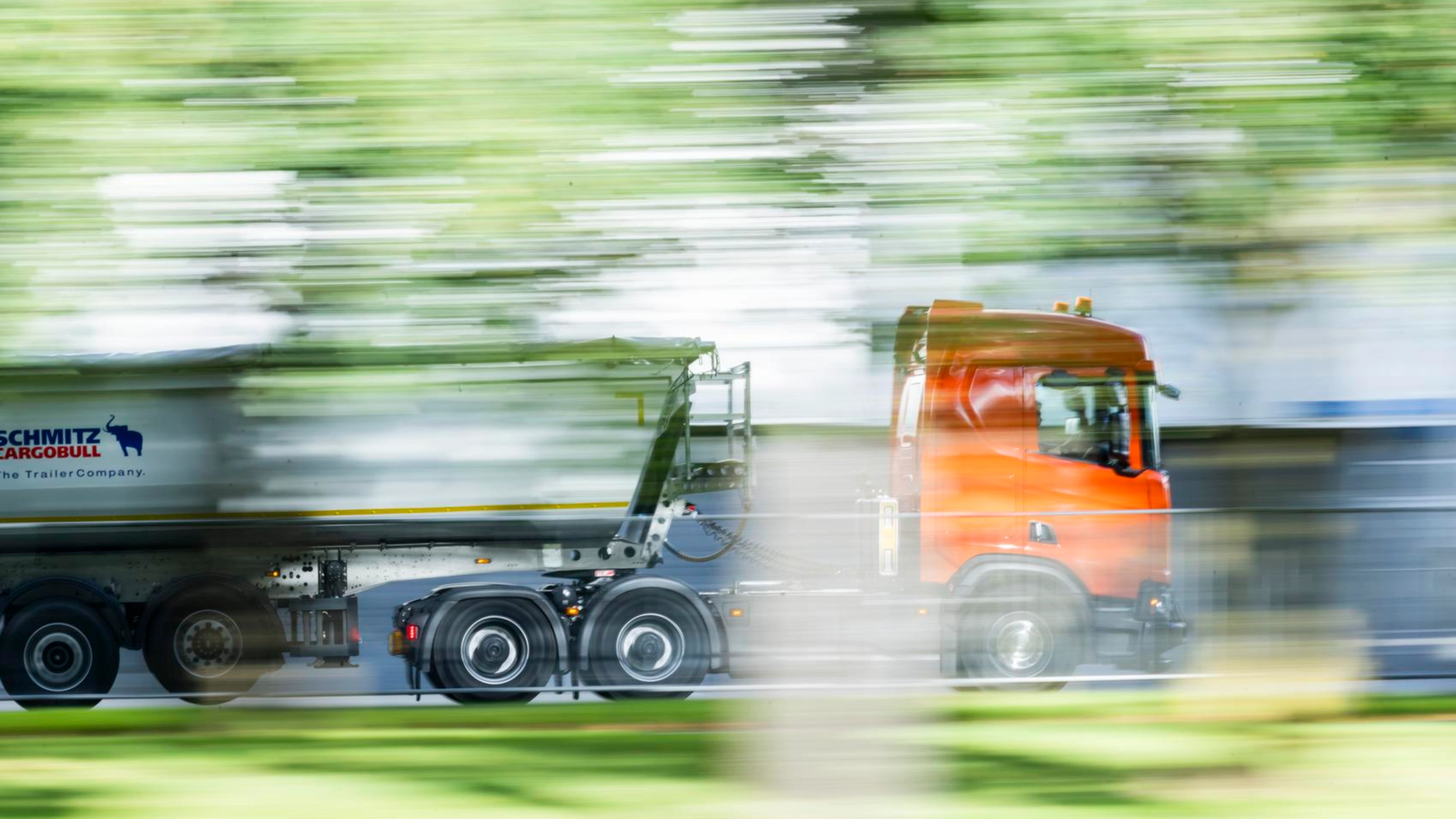 A truck with a trailer drives along a road surrounded by greenery.