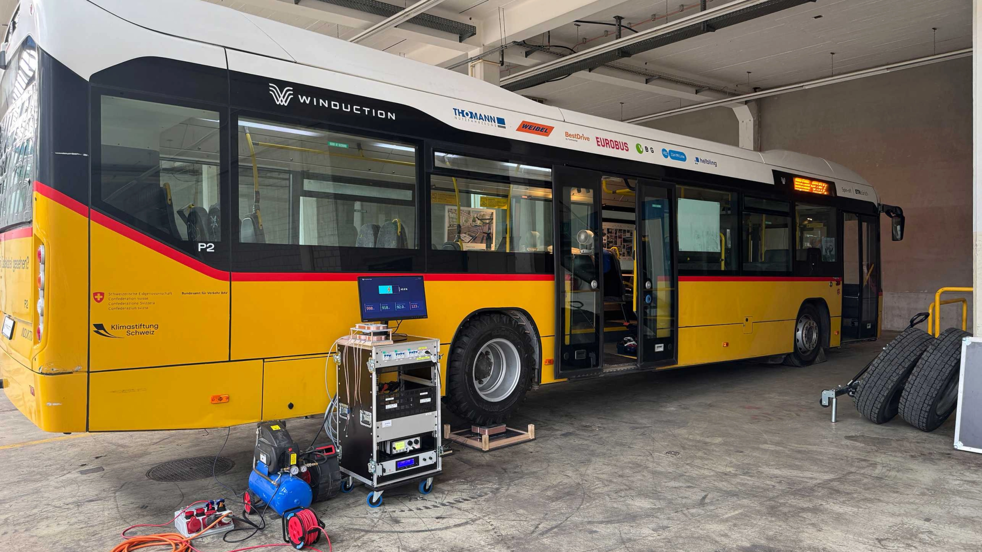 Yellow electric bus in a workshop with open doors and technical equipment in front of it.