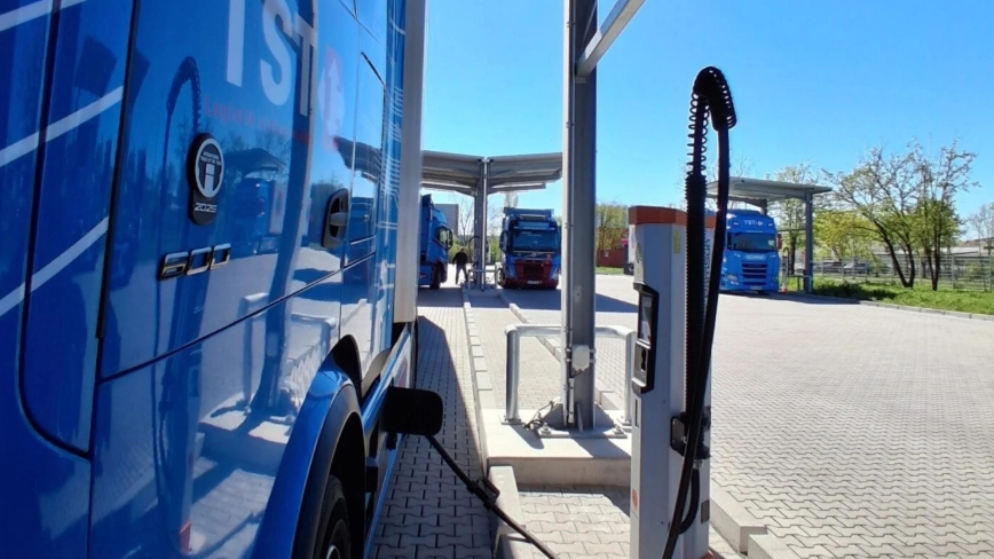 Blue electric truck connected to a charging station at a parking area.