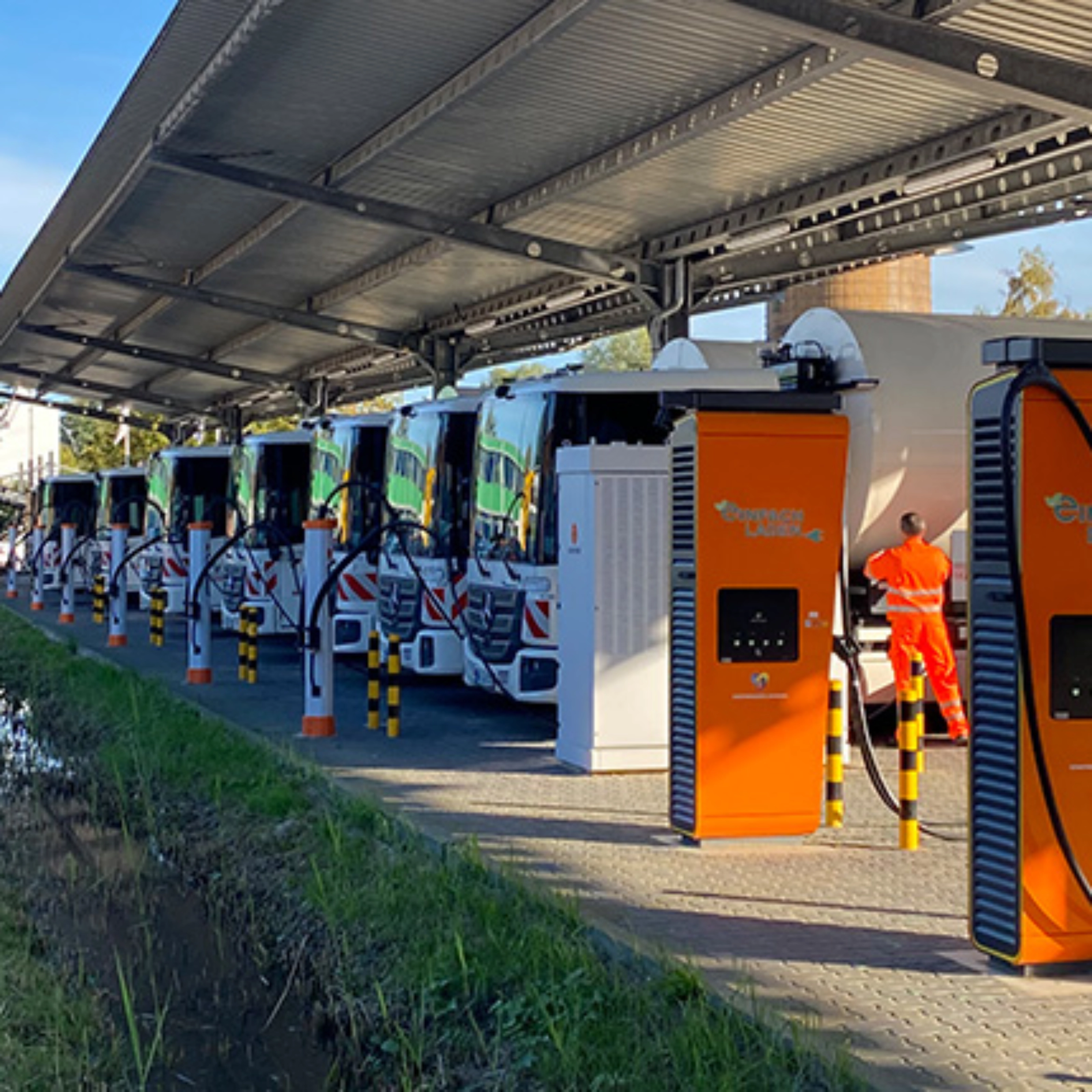 Orange charging stations beneath a solar canopy at a parking area with parked trucks.