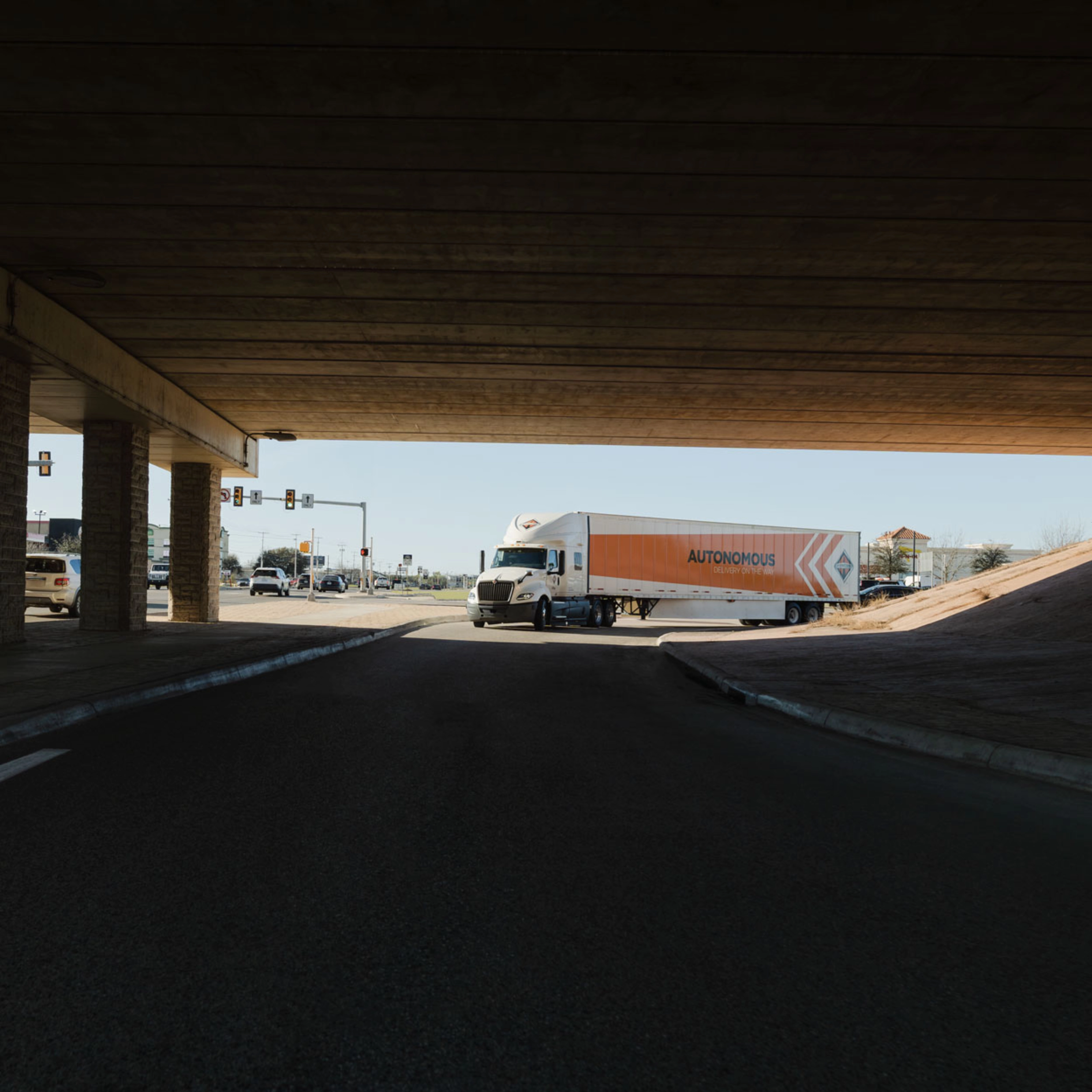 A self-driving truck with an "Autonomous" trailer drives through an intersection under a bridge.