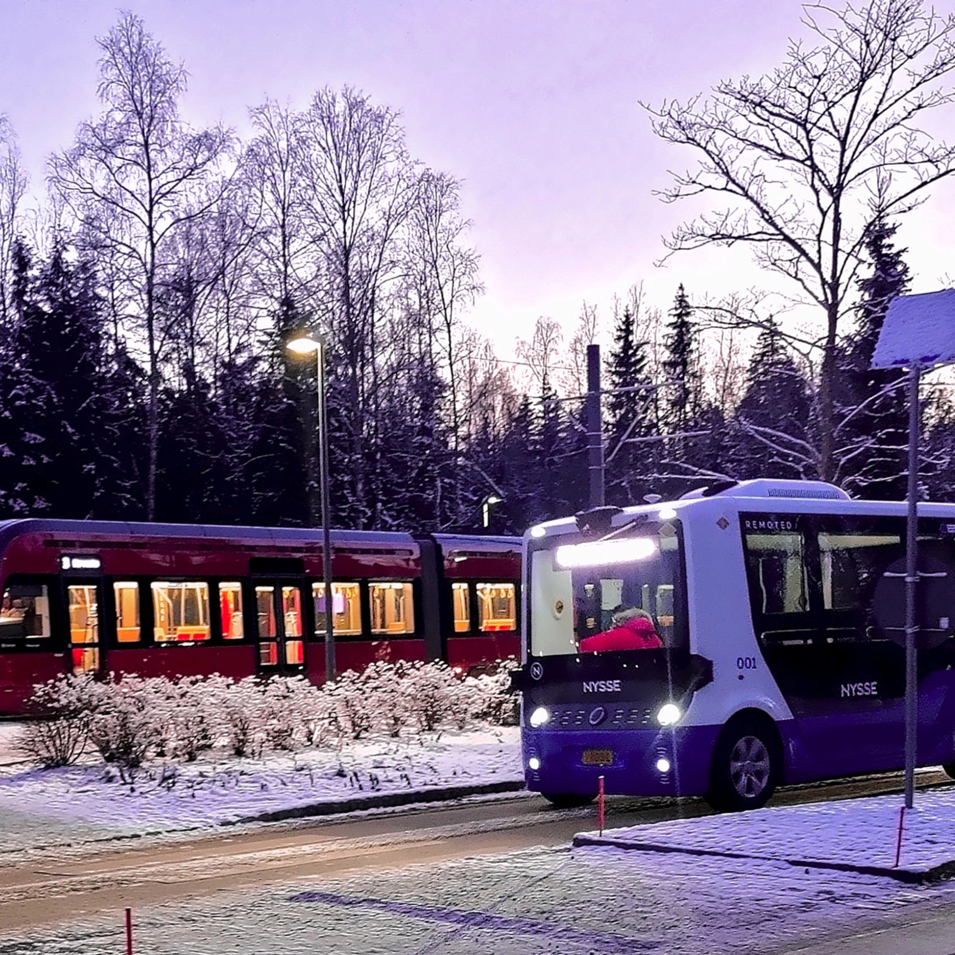 Autonomous shuttle bus driving beside a tram on a snowy street.