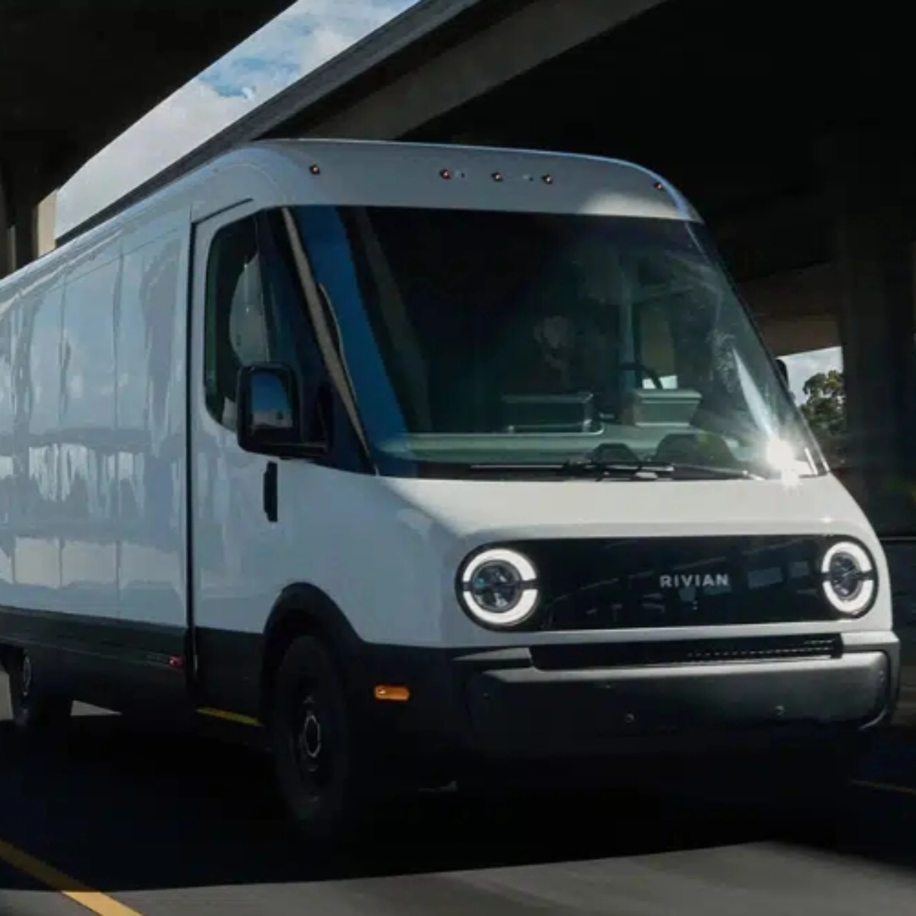 White Rivian electric delivery van driving on a city street under a highway bridge.
