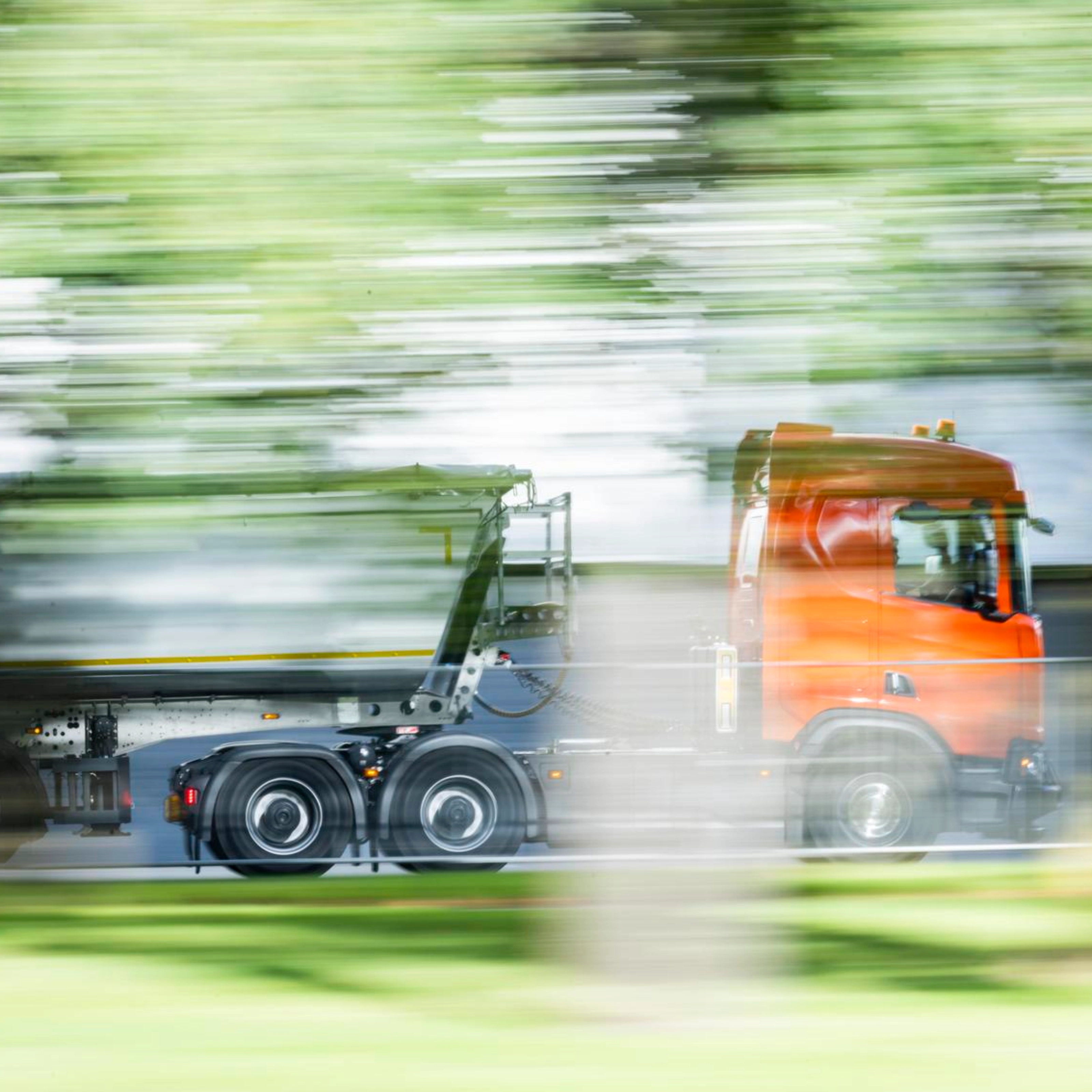 A truck with a trailer drives along a road surrounded by greenery.