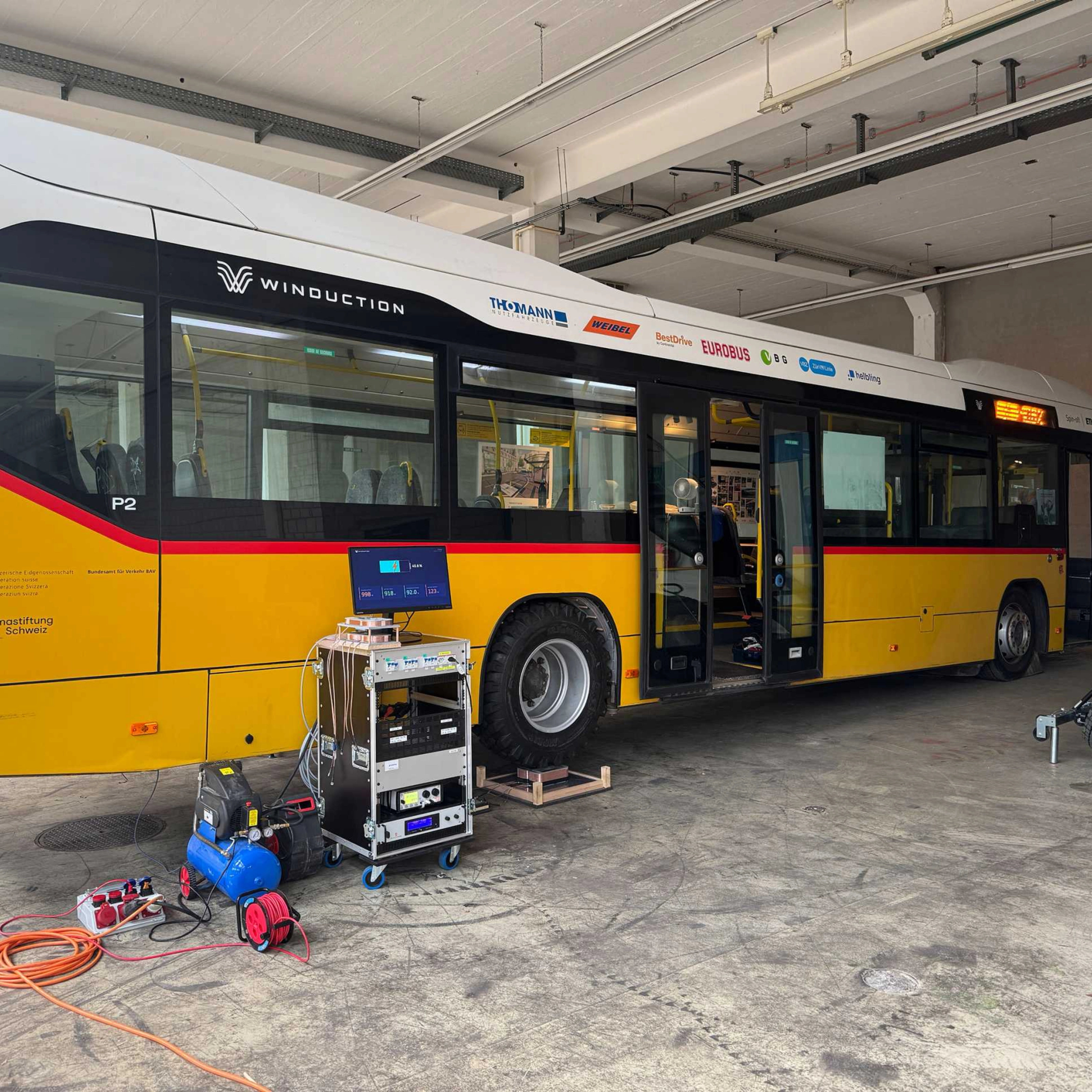 Yellow electric bus in a workshop with open doors and technical equipment in front of it.