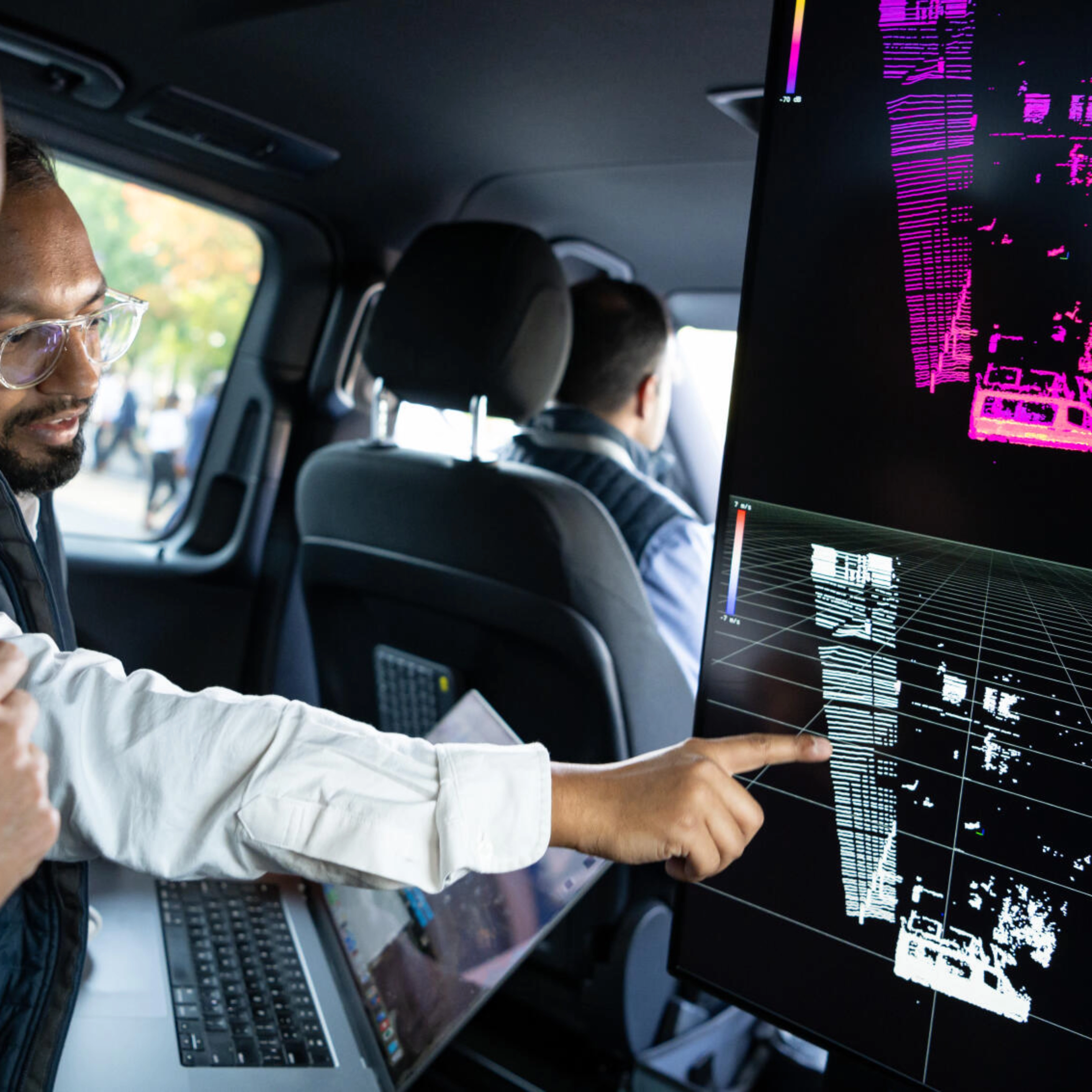 Two men in a car analyze 3D sensor data on a screen, likely testing autonomous driving tech.