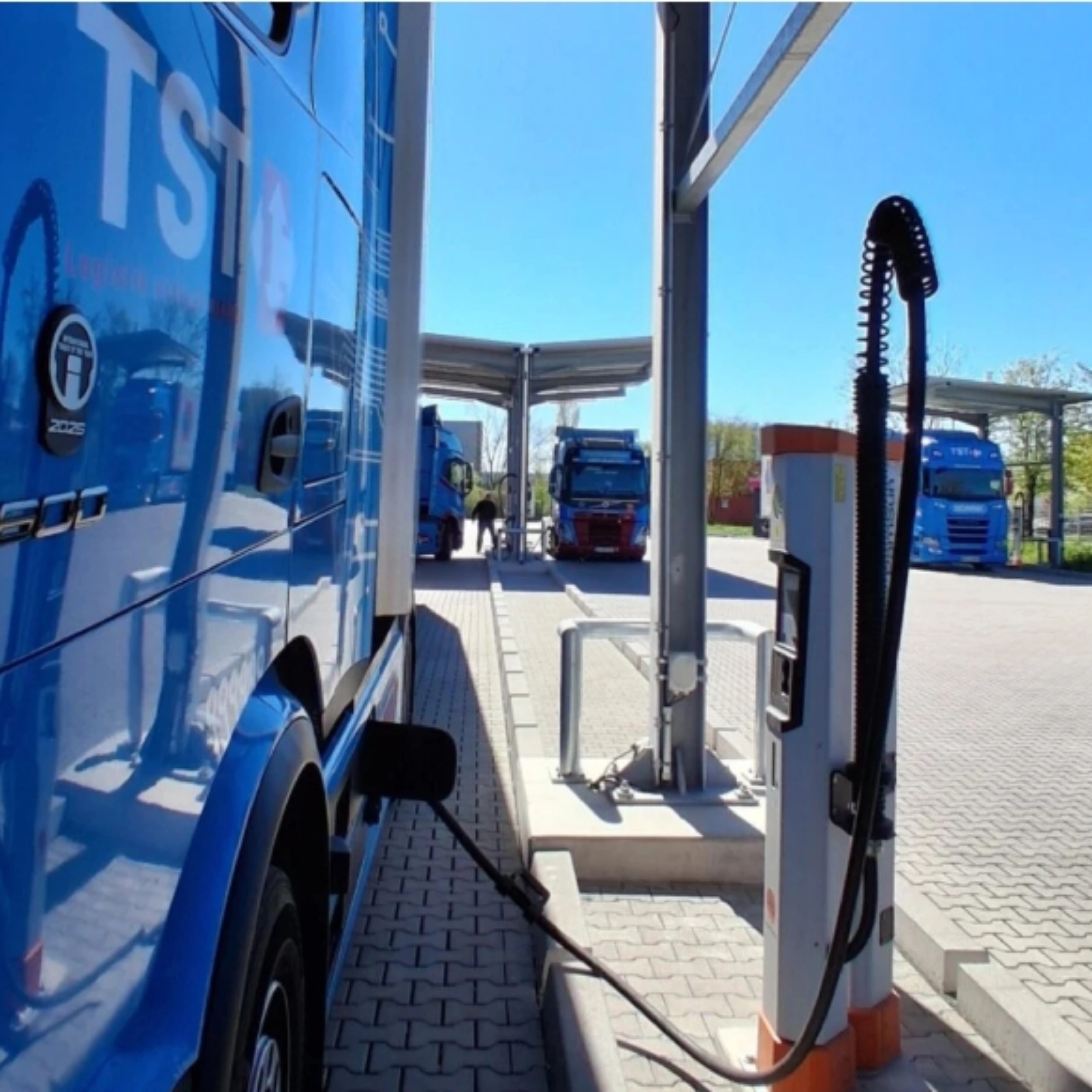 Blue electric truck connected to a charging station at a parking area.