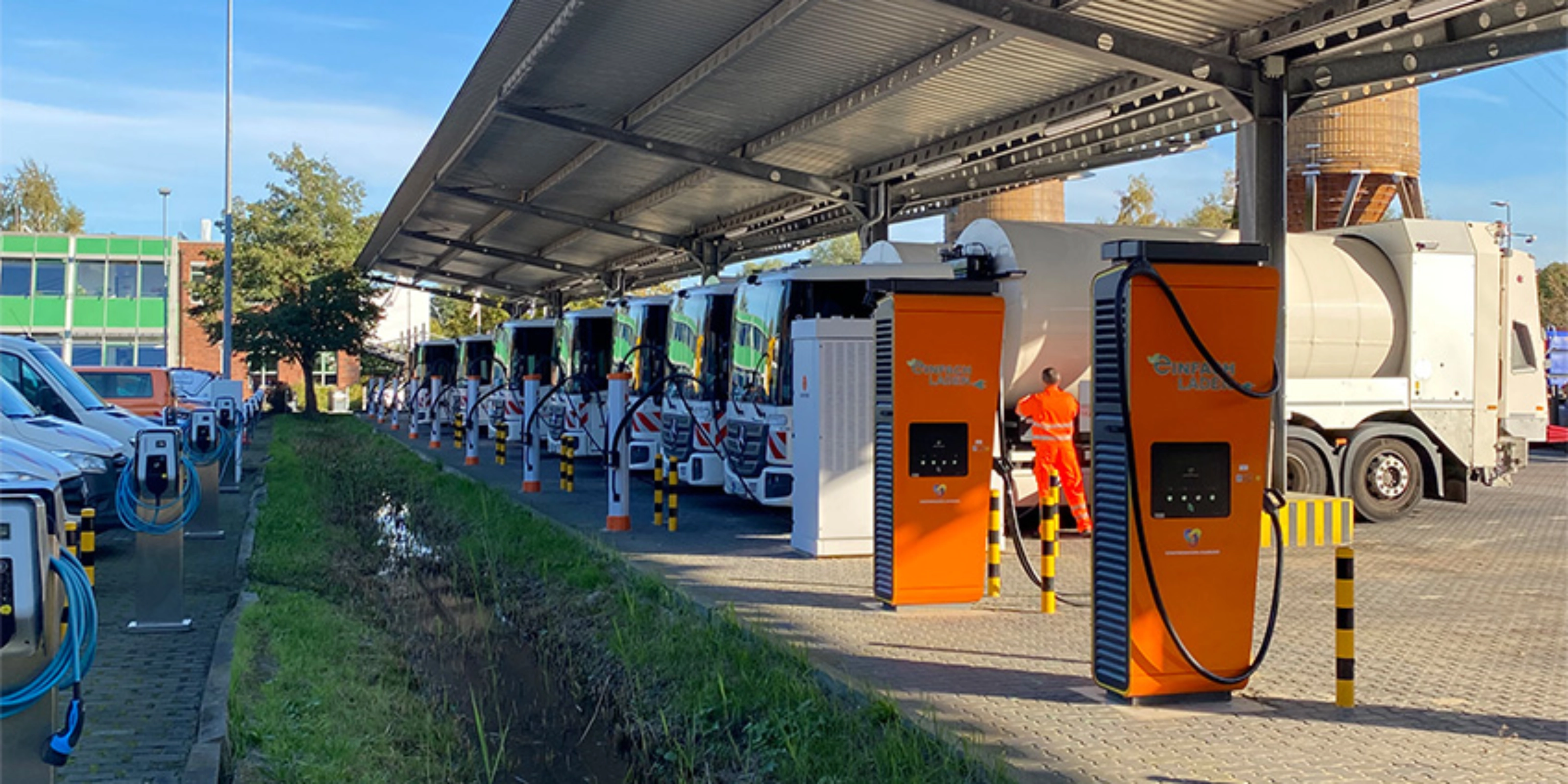 Orange charging stations beneath a solar canopy at a parking area with parked trucks.