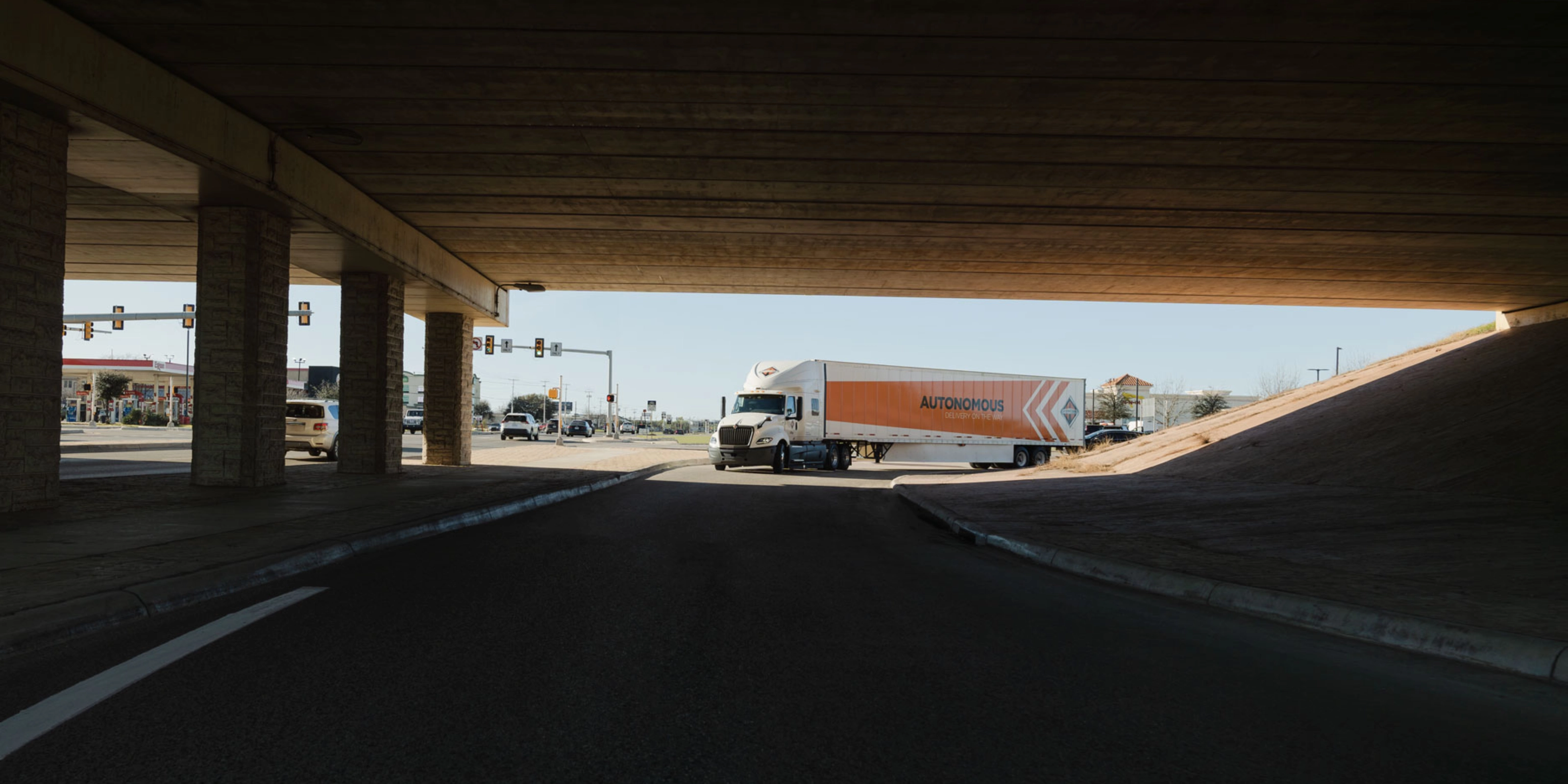 A self-driving truck with an "Autonomous" trailer drives through an intersection under a bridge.