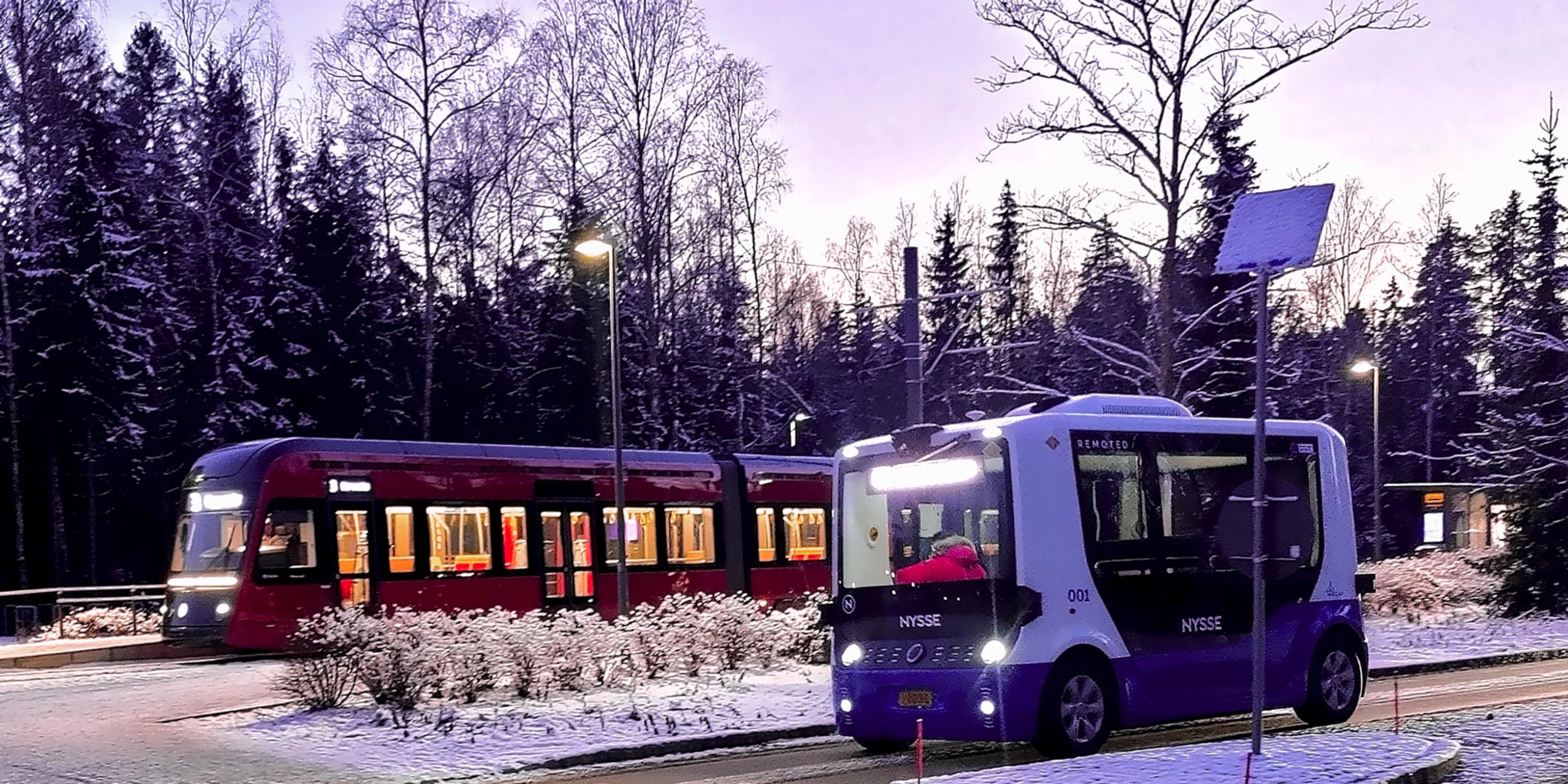Autonomous shuttle bus driving beside a tram on a snowy street.