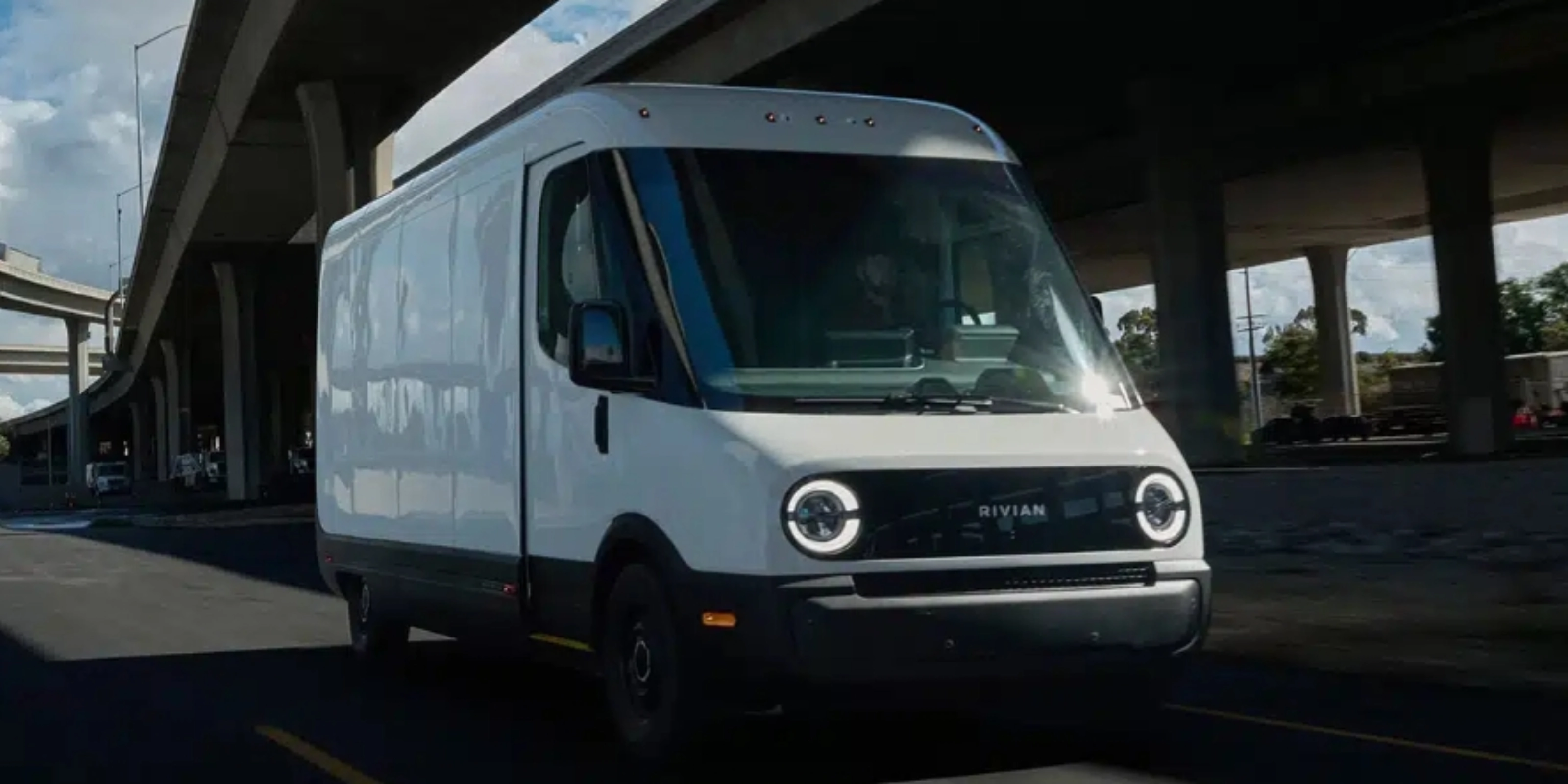 White Rivian electric delivery van driving on a city street under a highway bridge.