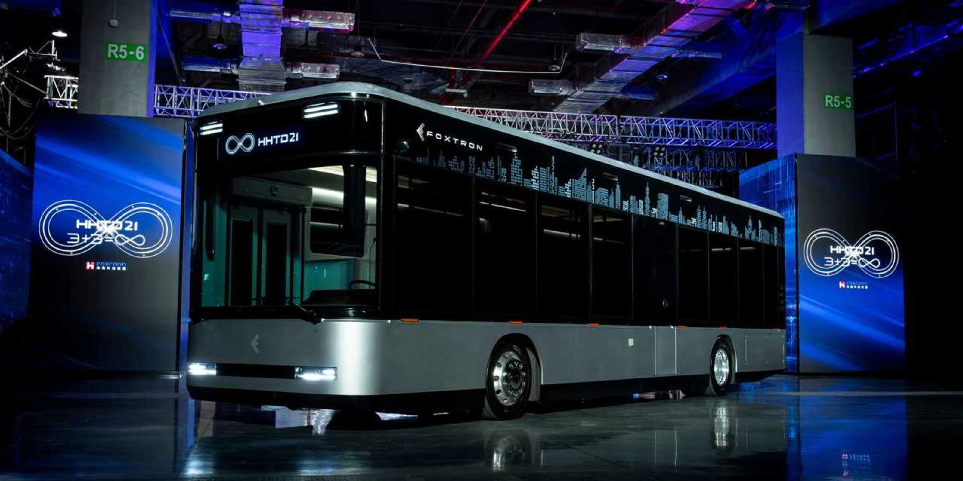 A modern electric city bus is showcased indoors with the logo "Foxtron" and digital skyline.
