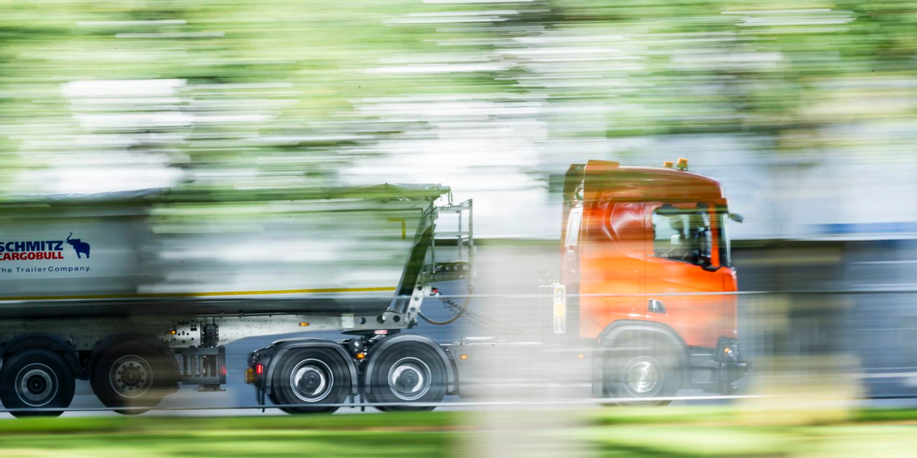 A truck with a trailer drives along a road surrounded by greenery.