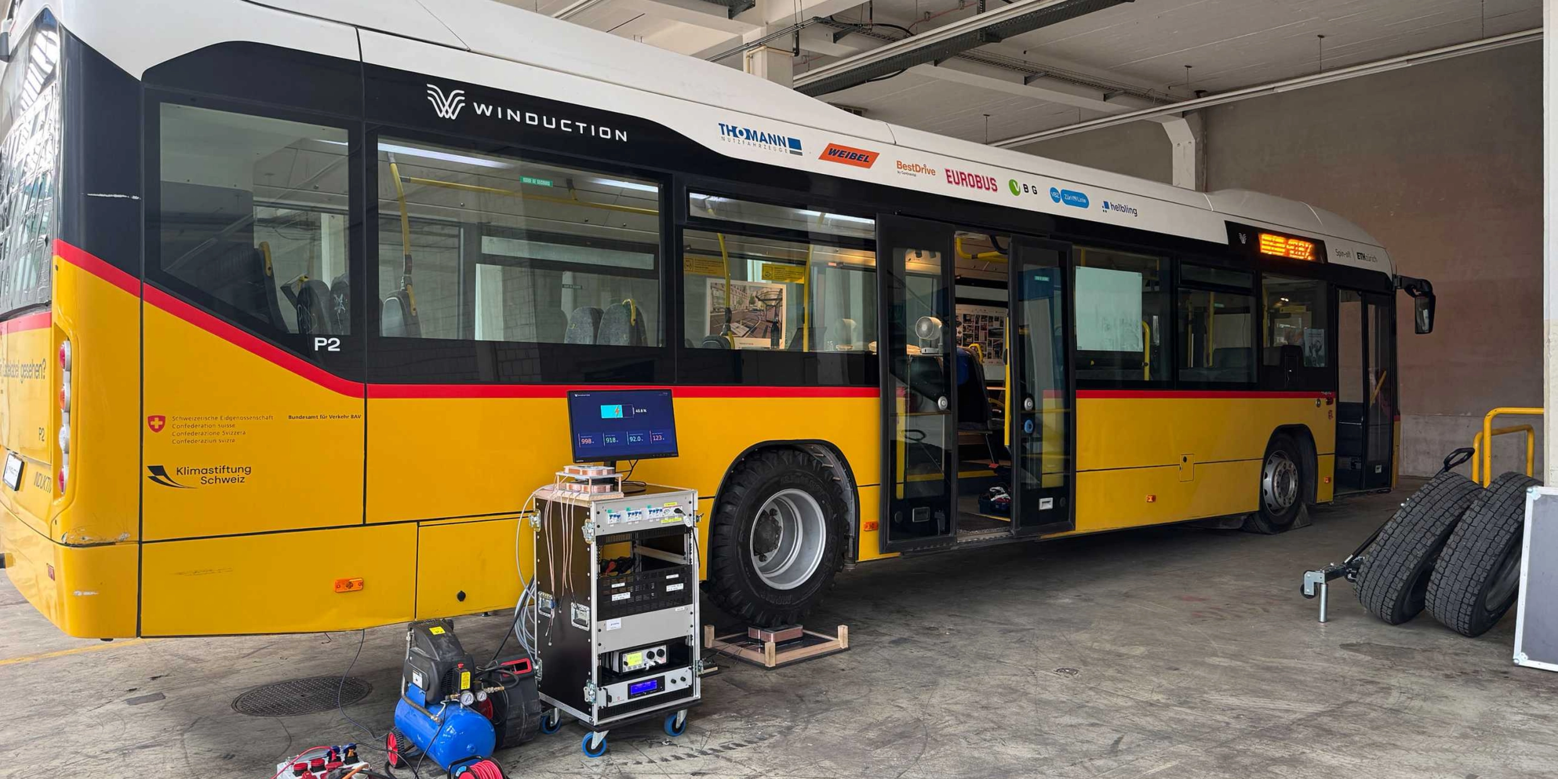 Yellow electric bus in a workshop with open doors and technical equipment in front of it.