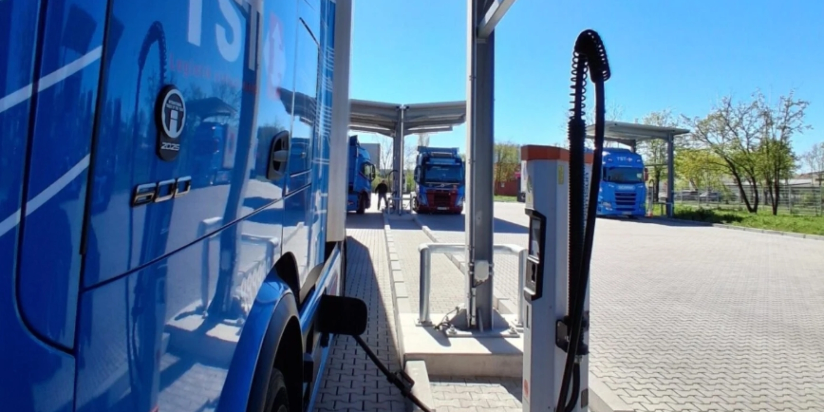 Blue electric truck connected to a charging station at a parking area.