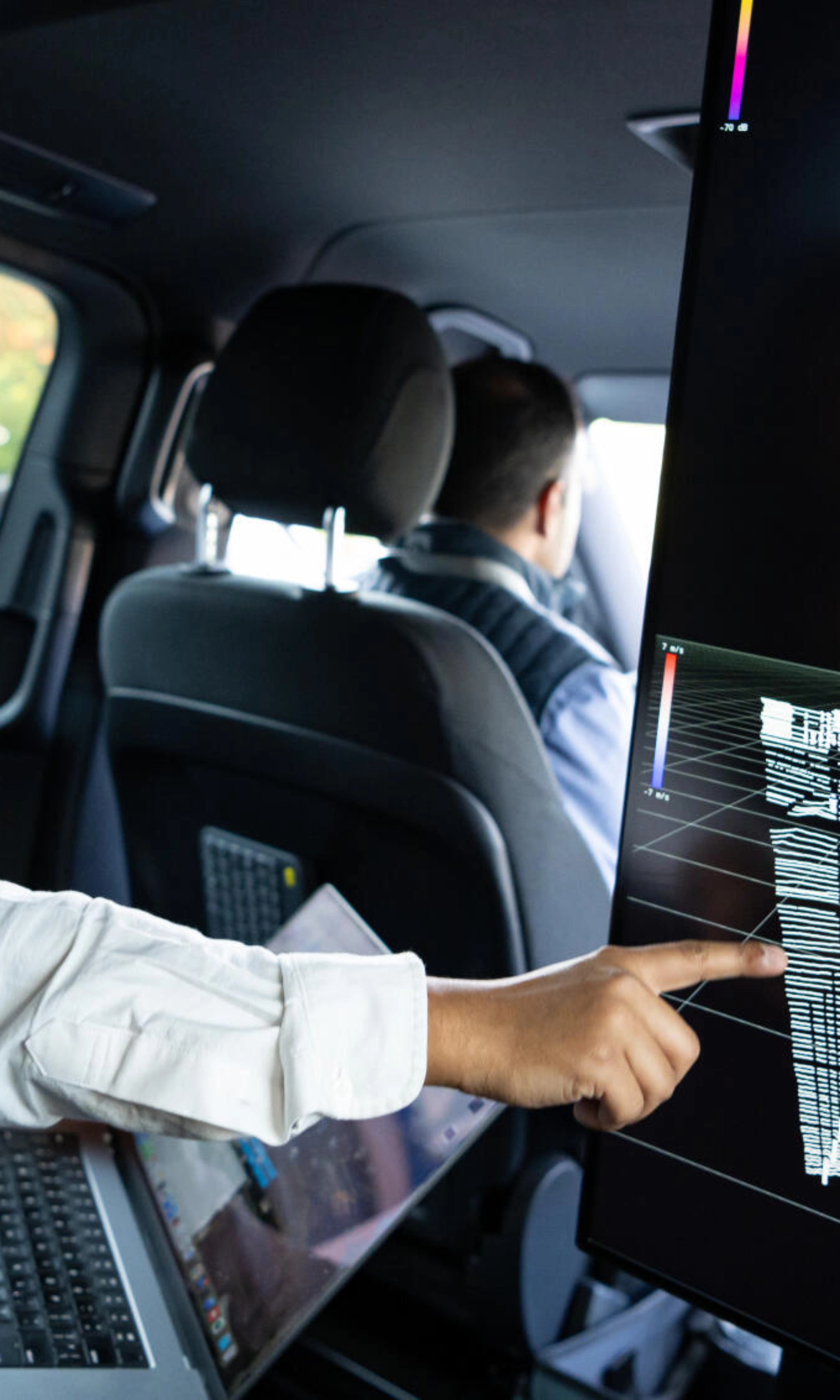 Two men in a car analyze 3D sensor data on a screen, likely testing autonomous driving tech.