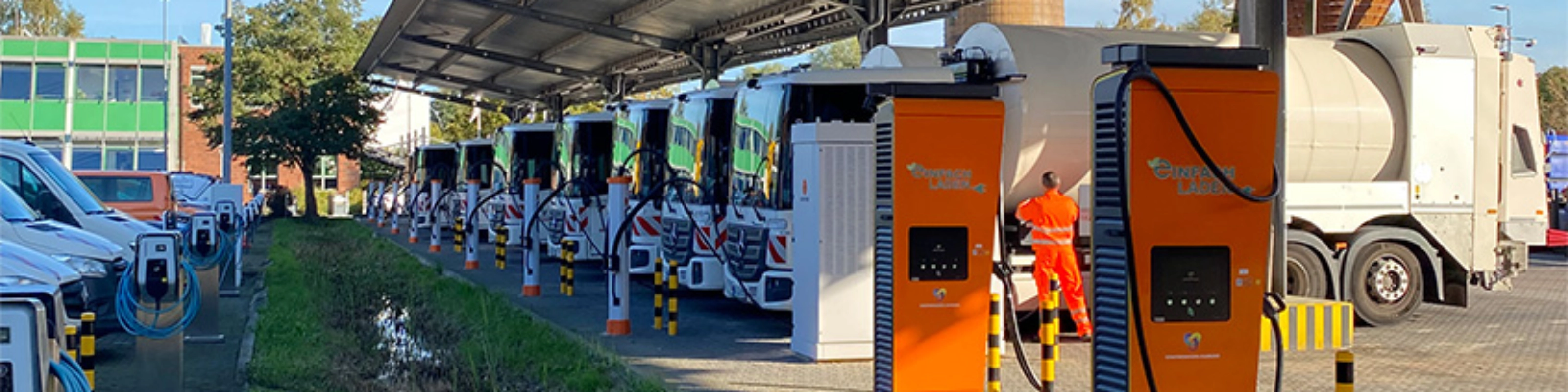 Orange charging stations beneath a solar canopy at a parking area with parked trucks.