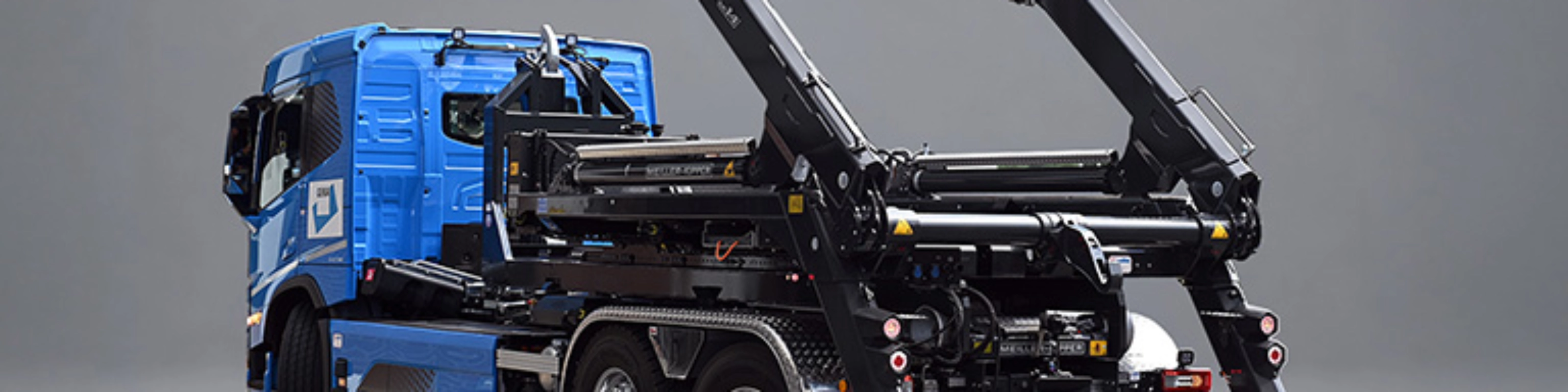 Blauer Lkw mit schwarzem Meiller-Hakenliftsystem und angehobenen Hubarmen in schräger Rückansicht.