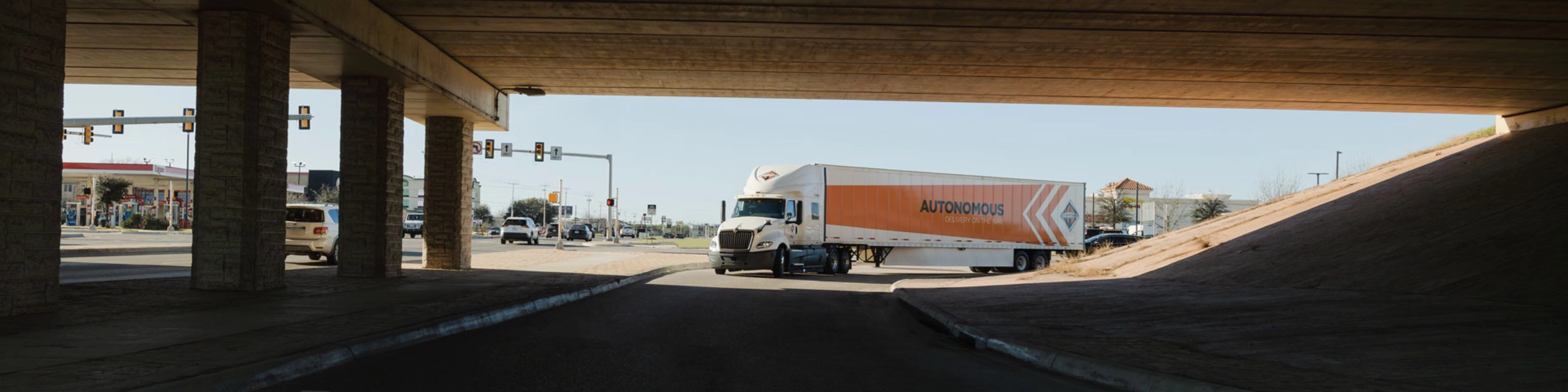 A self-driving truck with an "Autonomous" trailer drives through an intersection under a bridge.