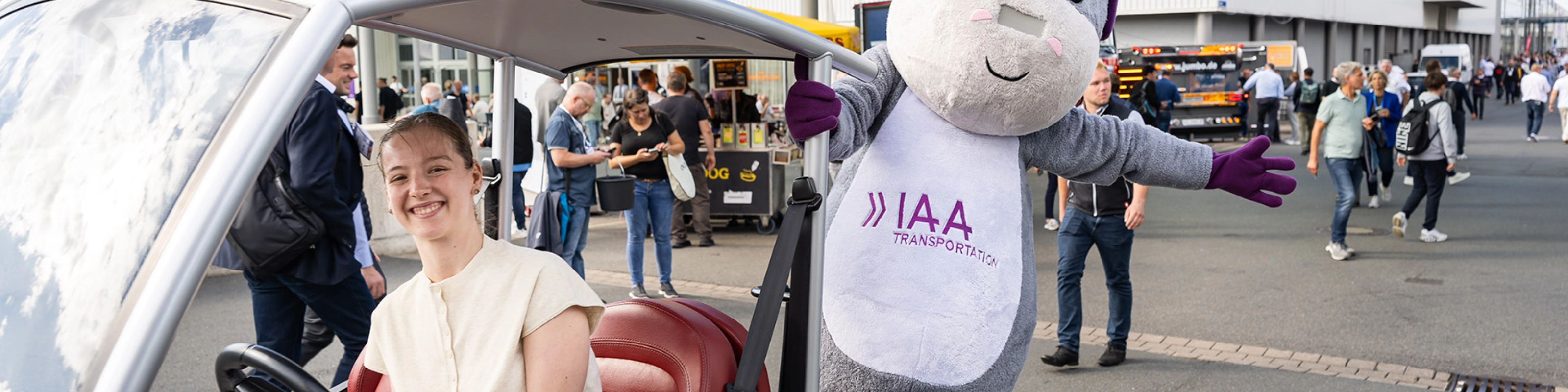 Frau in einem IAA-Fahrzeug neben dem IAA Transportation Maskottchen auf der Messe.