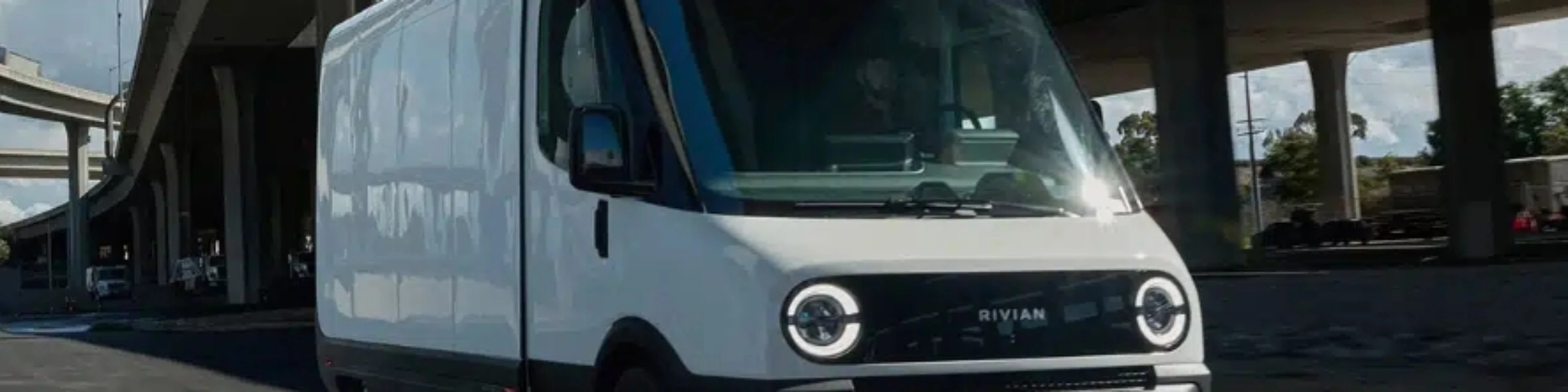 White Rivian electric delivery van driving on a city street under a highway bridge.
