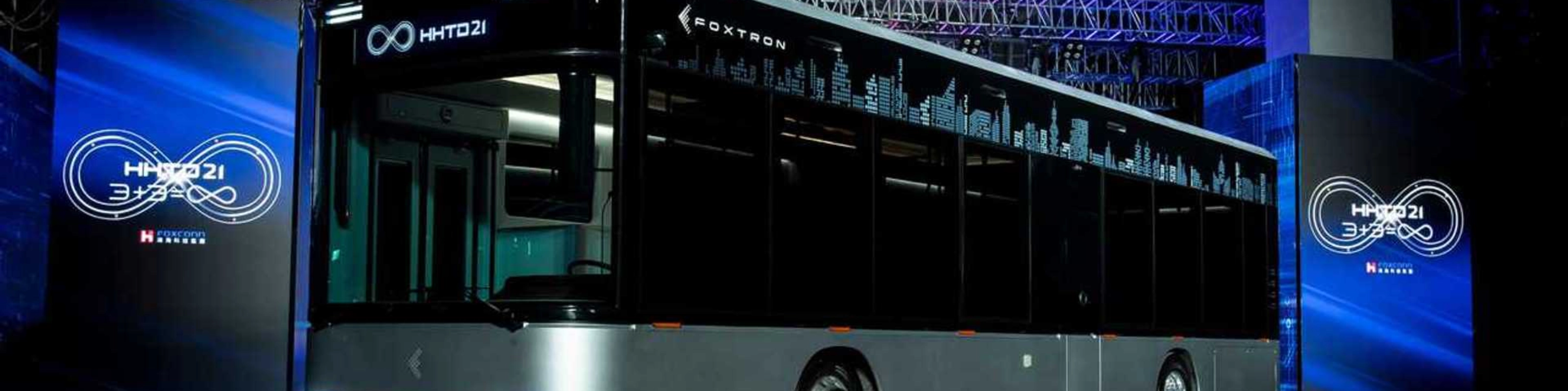 A modern electric city bus is showcased indoors with the logo "Foxtron" and digital skyline.