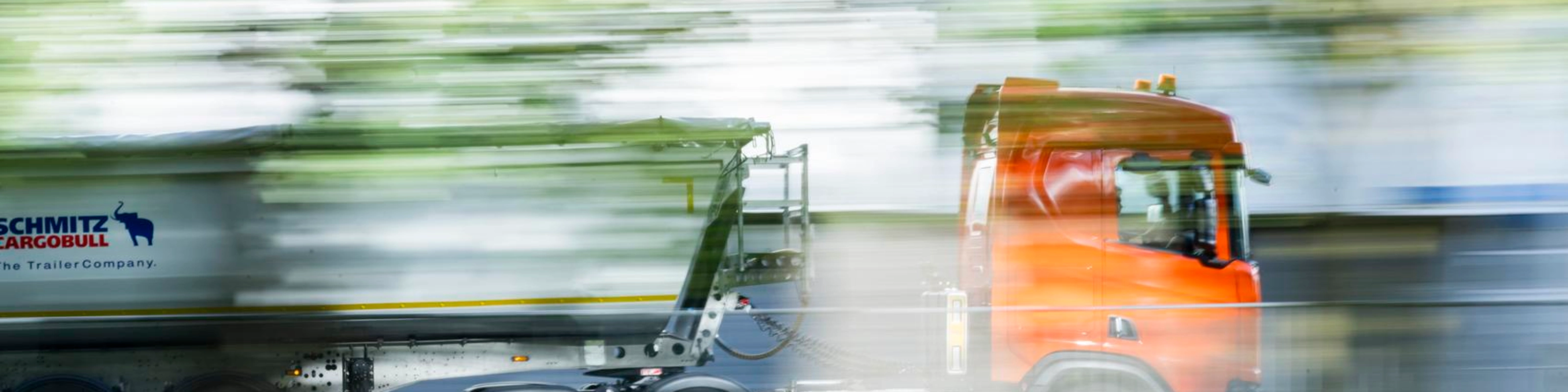 A truck with a trailer drives along a road surrounded by greenery.