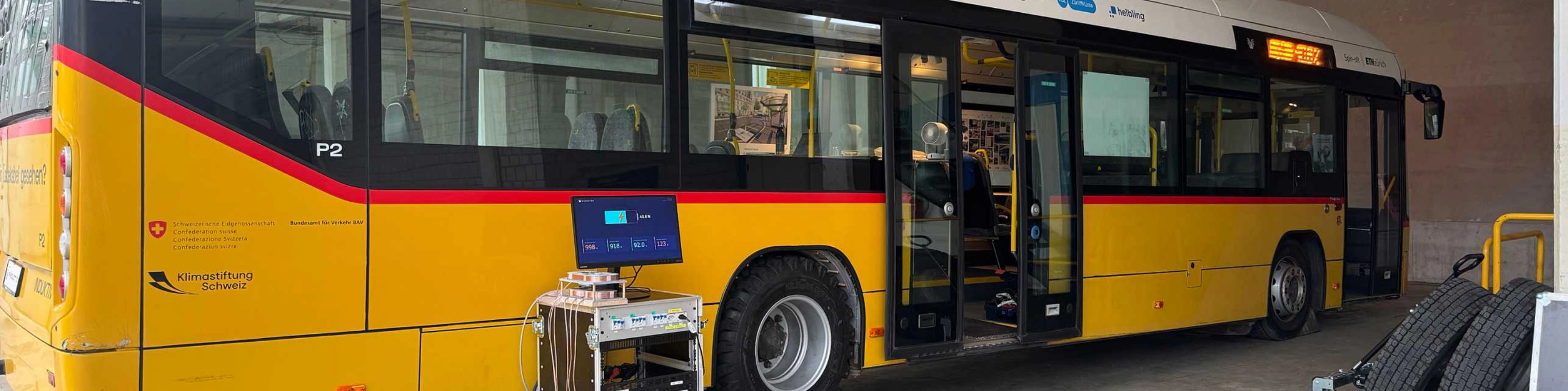 Yellow electric bus in a workshop with open doors and technical equipment in front of it.