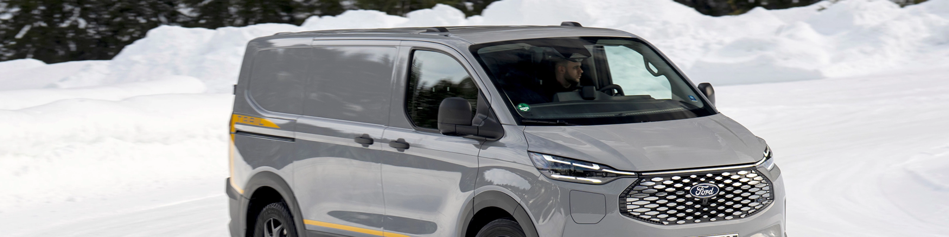 Grey Ford electric van driving on a snowy road with forest in the background.