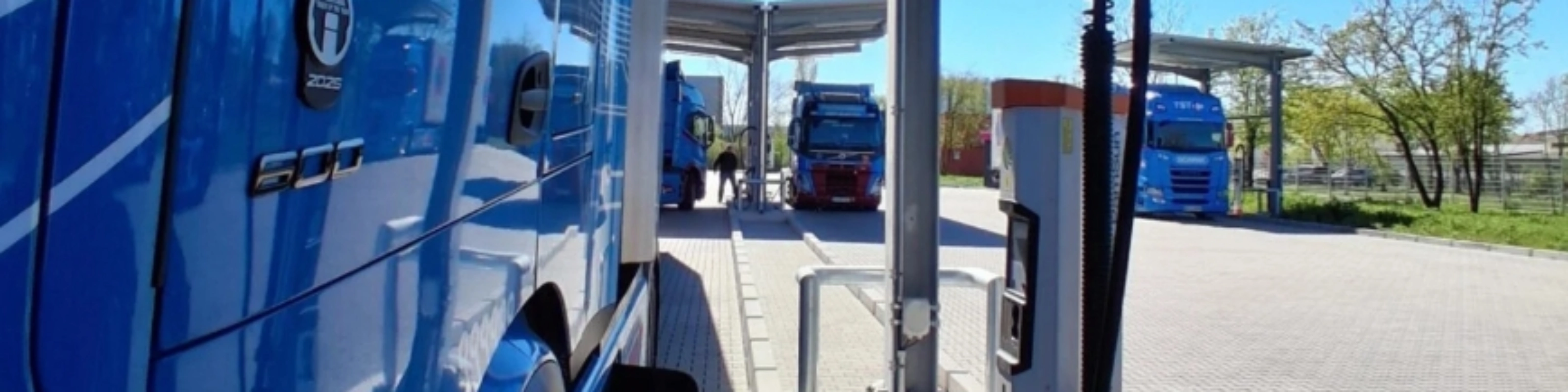 Blue electric truck connected to a charging station at a parking area.