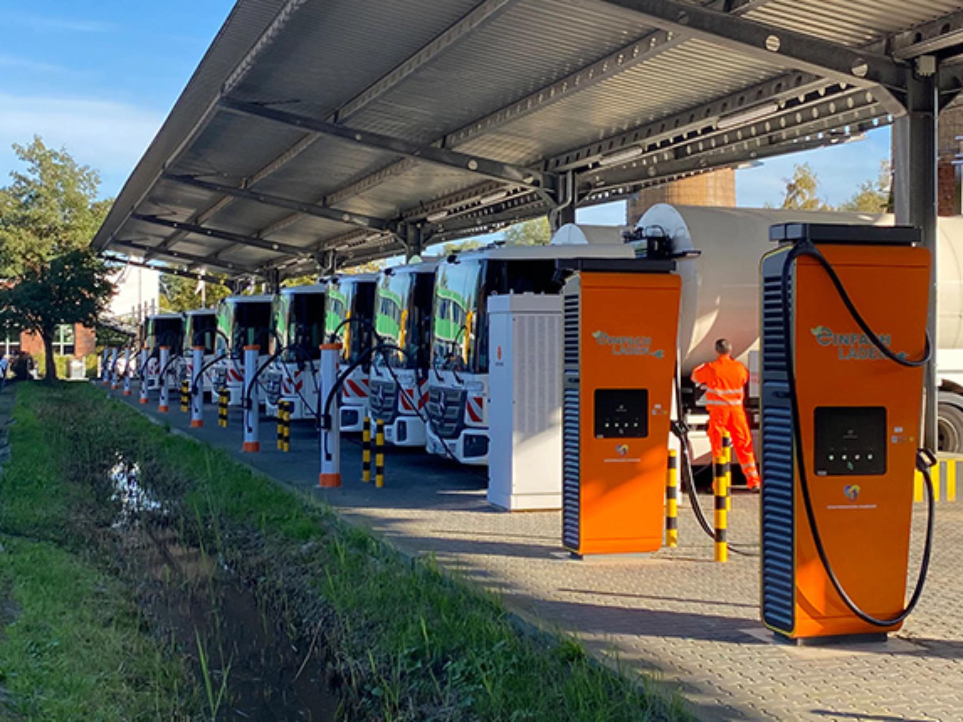 Orange charging stations beneath a solar canopy at a parking area with parked trucks.