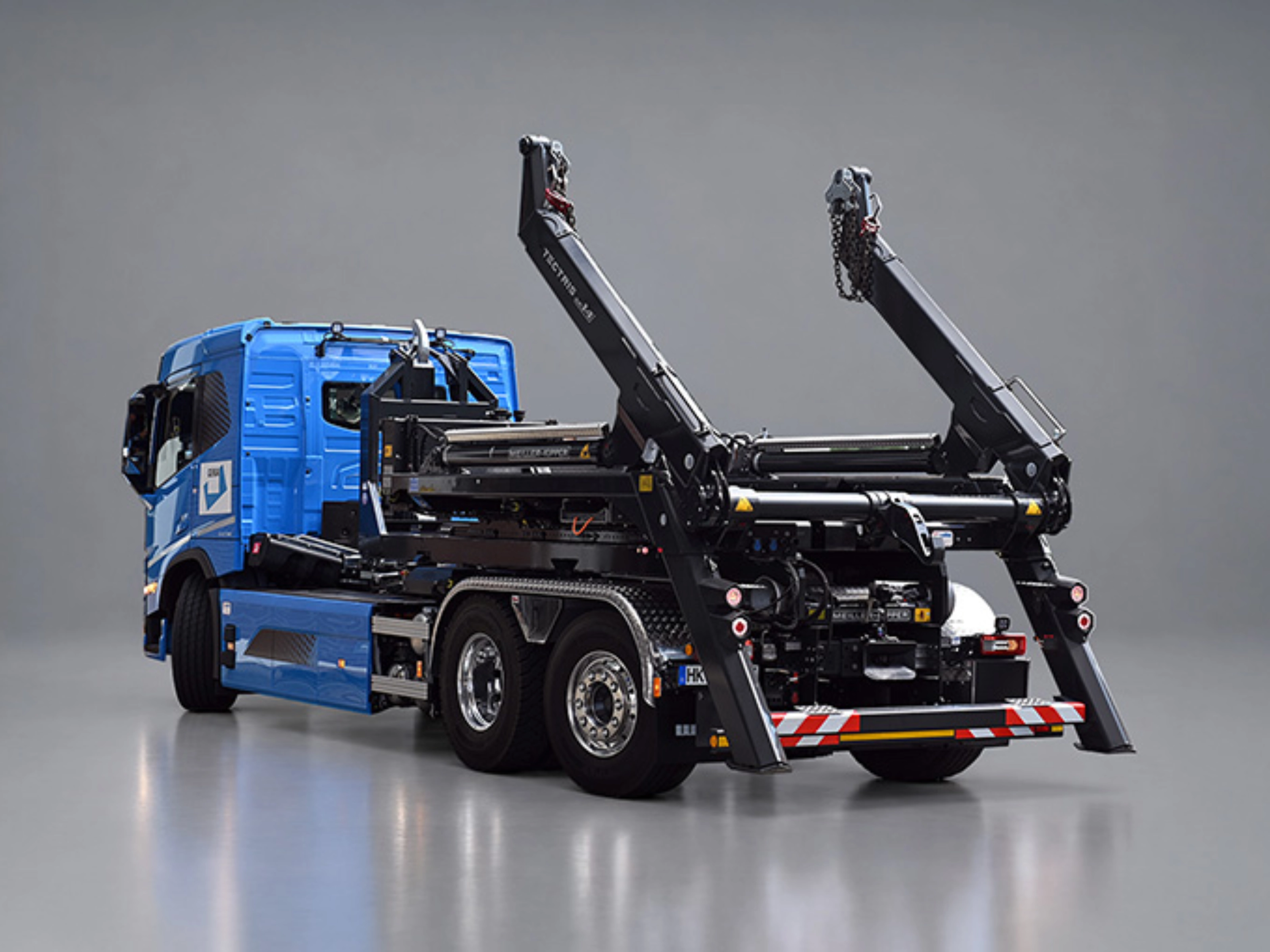 Blauer Lkw mit schwarzem Meiller-Hakenliftsystem und angehobenen Hubarmen in schräger Rückansicht.