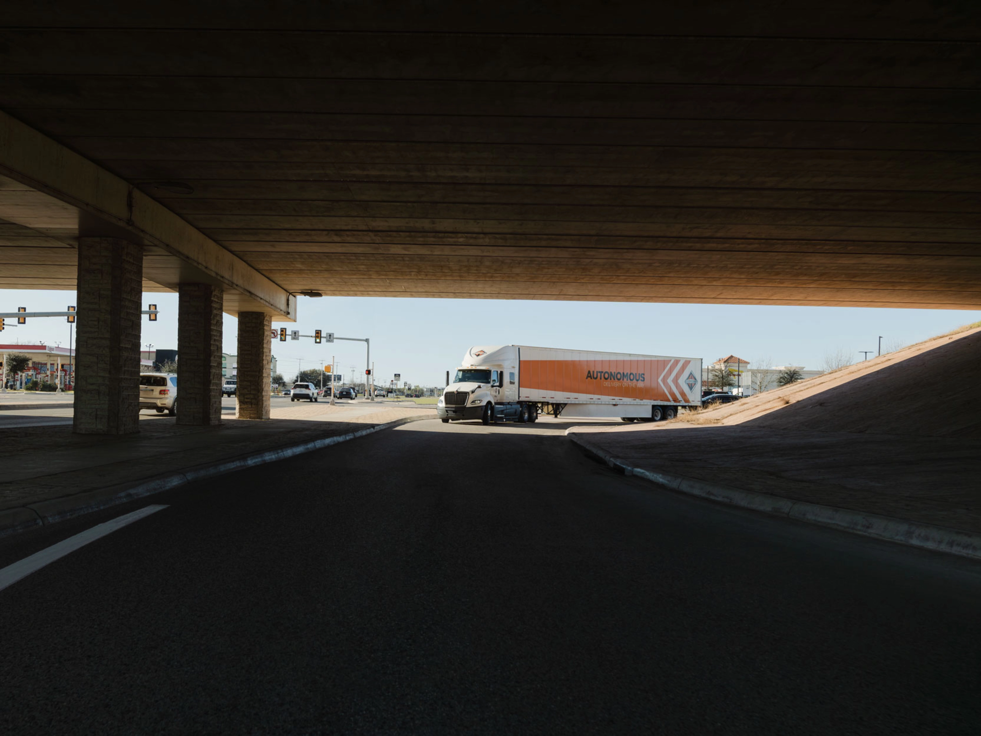 A self-driving truck with an "Autonomous" trailer drives through an intersection under a bridge.