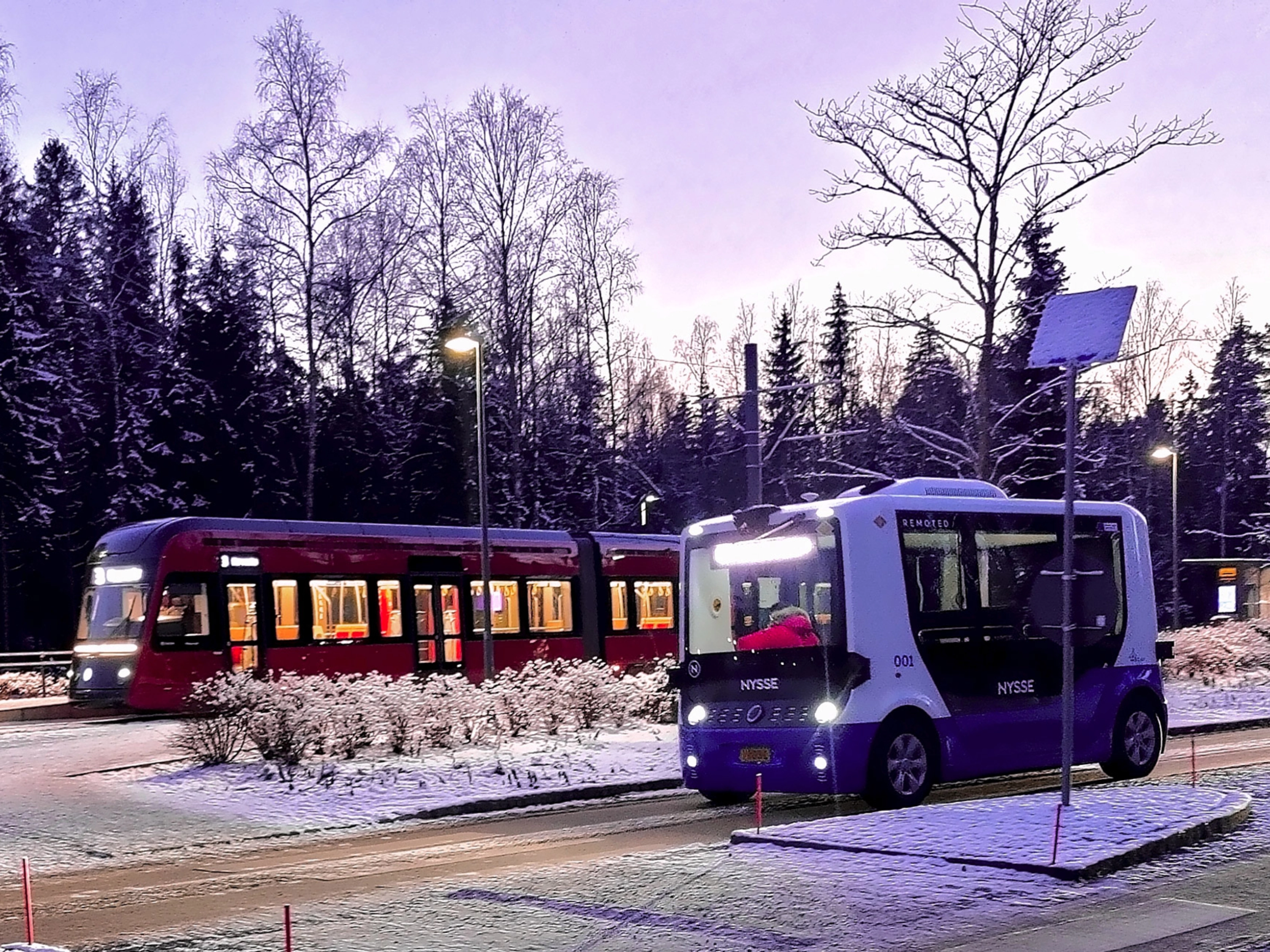 Autonomous shuttle bus driving beside a tram on a snowy street.