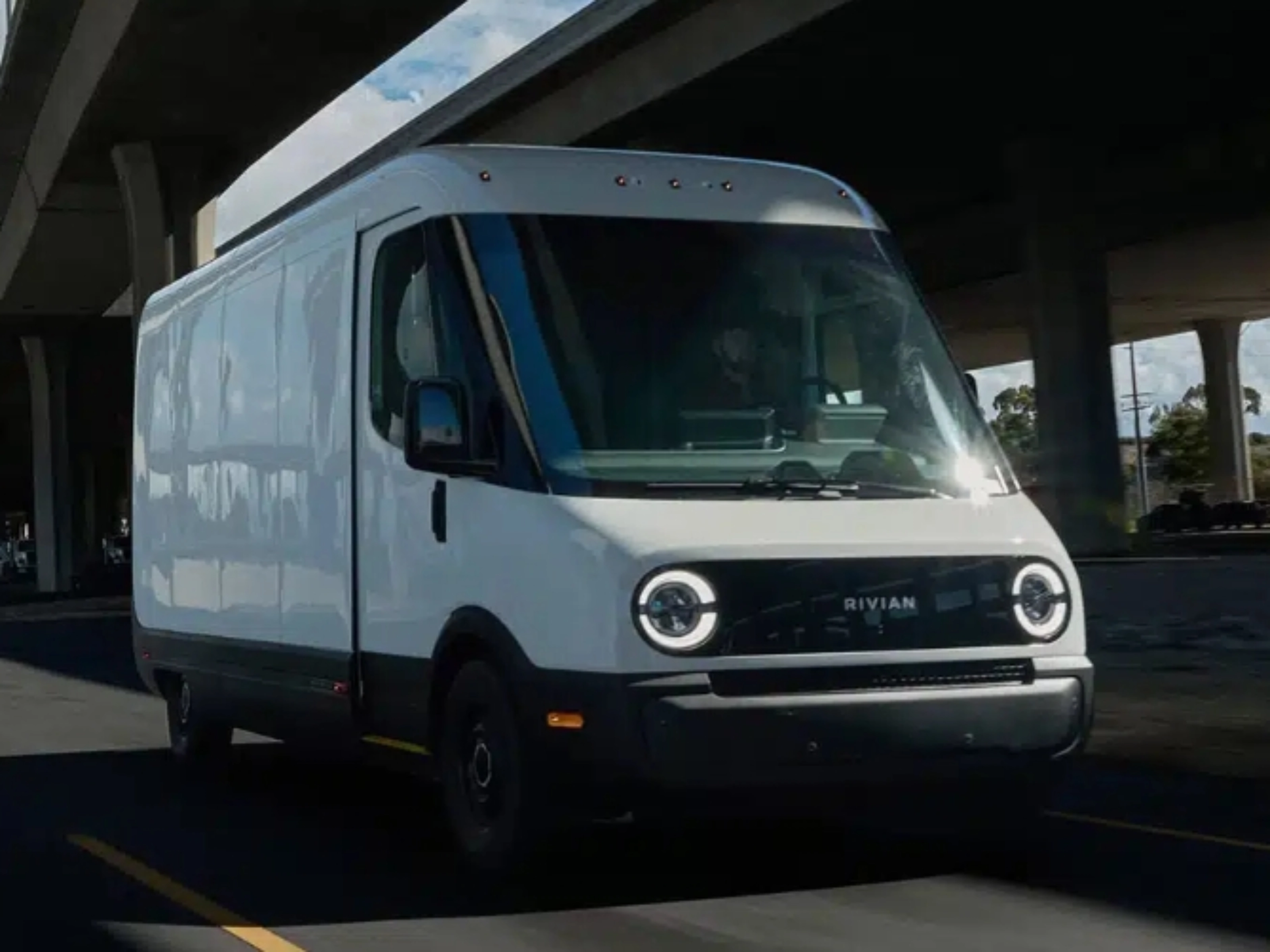 White Rivian electric delivery van driving on a city street under a highway bridge.